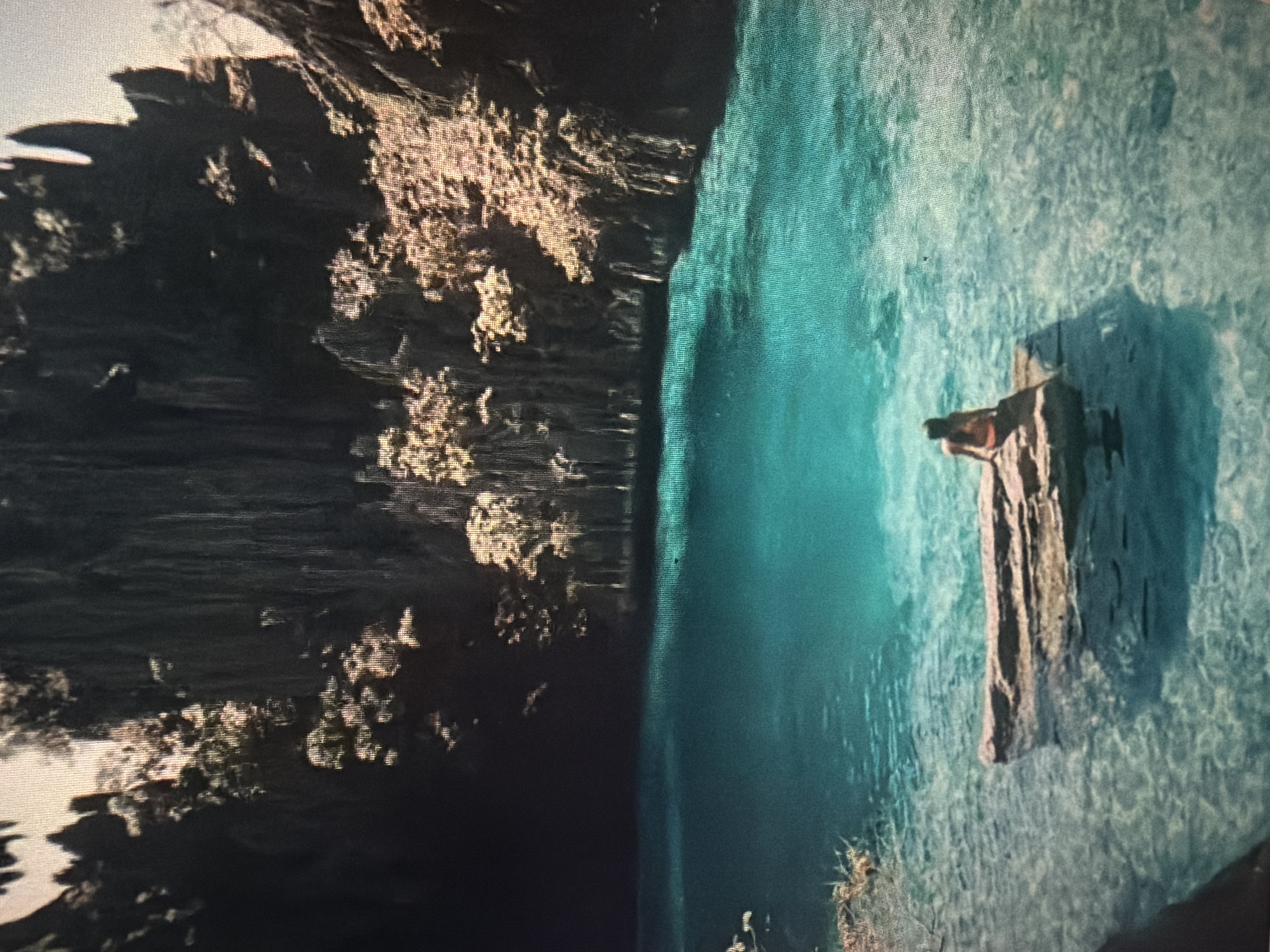 A person sits on a rock in a clear turquoise lagoon surrounded by steep, rocky cliffs covered in vegetation. The person is seen from behind and is wearing an orange top. The water is shallow enough to see the rocks and sand on the bottom. The scene appears to be a tropical paradise. The provided location information, "Baan Mae Nam, Thailand," is not visually verifiable from the image alone.