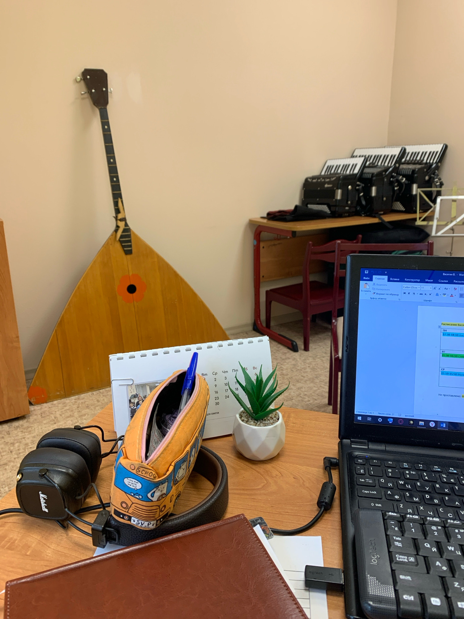 The image depicts a desk in what appears to be a music classroom or practice space. Dominating the background is a large, wooden balalaika leaning against a light-colored wall. To the right, three accordions are neatly arranged on a wooden table with red metal legs, suggesting a focus on musical instruments.
In the foreground, the desk surface is covered with various items. A black laptop, partially open and displaying a document, is prominent. To the left of the laptop are a pair of black Marshall headphones and a brightly colored pencil case adorned with cartoon characters of a school bus and animals. A blue pen is visible extending from the pencil case. A small artificial potted plant sits to the right of the pencil case, adding a touch of greenery. A desk calendar with Cyrillic lettering is also present, with some dates visible. Underneath the laptop, a leather-bound notebook or planner is partially visible. A logitech USB device is plugged into the laptop, and another USB cable runs from the laptop to a black adapter. The overall environment is a cluttered but organized workspace, likely for musical study or performance.
Based on available information, Katunino, Russia, is known for its aviation technical school and associated cultural activities, making it plausible that such a music-related space could exist there.