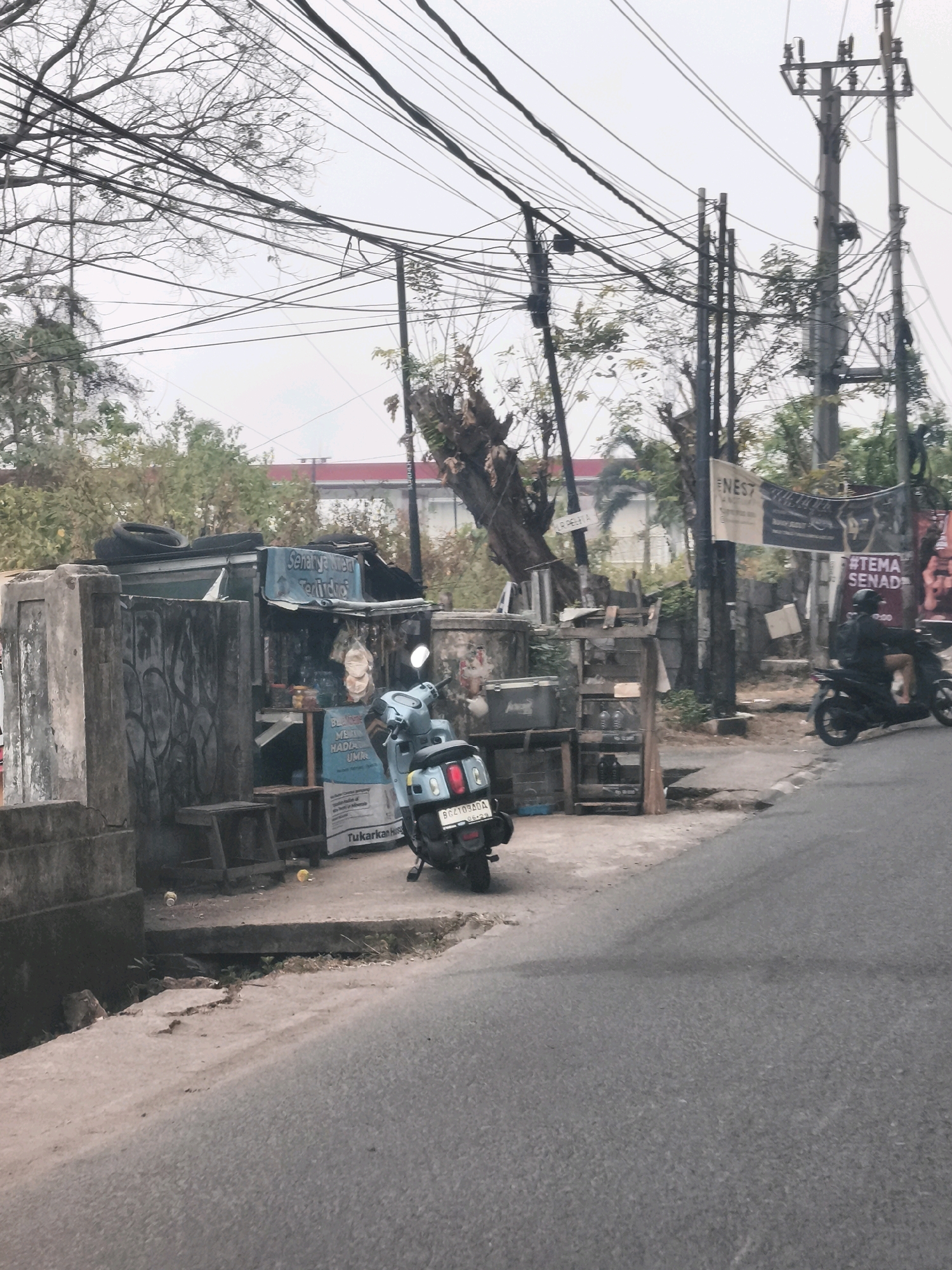 The image depicts a street scene in Palembang, Indonesia, likely in a more modest urban area characterized by utility poles and overhead wires, a common sight in the city. In the foreground, a paved road runs diagonally across the frame, with a rough concrete sidewalk bordering it. On the sidewalk, a small roadside stall is set up, selling various items, including what appear to be drinks in bottles. A light blue scooter is parked in front of the stall. The stall itself is constructed from a mix of corrugated metal and wood, with a tarp acting as a roof. Signs are visible, some in Indonesian, advertising products or services. A person wearing dark clothing is seated on a darker-colored scooter further down the street, also on the right side of the road, facing away from the camera. Behind the stall, trees and foliage are visible, interspersed with more utility poles. In the background, a building with a red roof can be seen, suggesting a residential or commercial area. The overall atmosphere is overcast, with a hazy sky. The scene conveys a sense of everyday life on a street in Palembang, with commercial activity taking place along a busy thoroughfare.