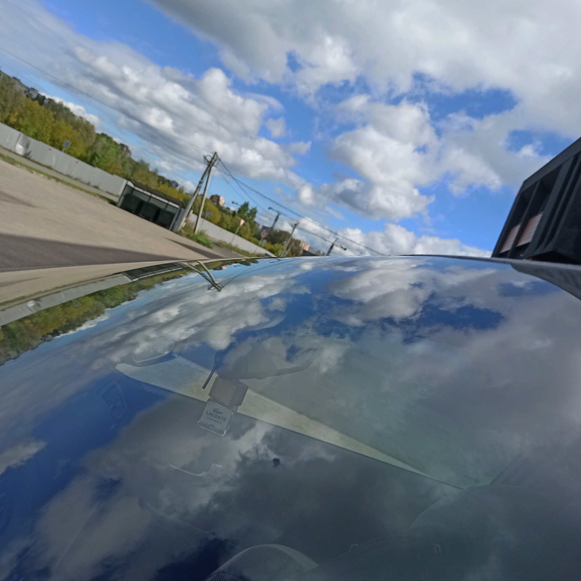 The image shows a view from inside a car, looking outwards and upwards. The windshield is reflective and shows a mirrored image of the sky, clouds, and trees. In the reflection on the windshield, a small rectangular air freshener with the brand "LACOSTE" is visible hanging from the rearview mirror. Outside the car, a grey asphalt road is visible, bordered by a light-colored fence. Beyond the fence, there are trees with green and yellow foliage, indicating it might be autumn. A utility pole with wires stretches across the scene. In the distance, there are buildings and more trees. The sky is blue with scattered white clouds. The scene appears to be in an urban or suburban area on a bright, partly cloudy day. The location is identified as Nizhny Novgorod, Russia.