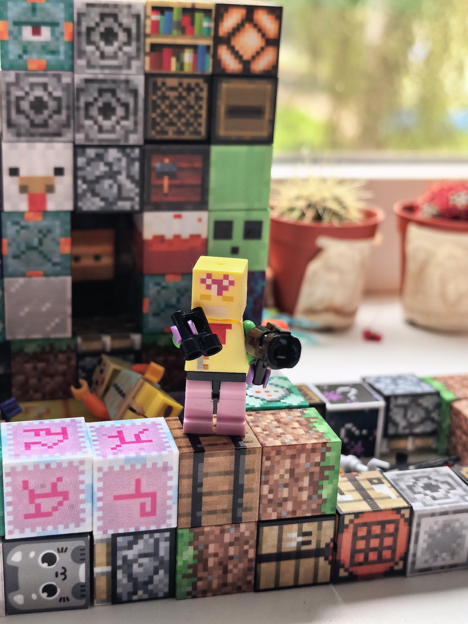 A close-up, eye-level shot captures a Lego minifigure with a yellow torso, pink legs, and a yellow head with pink facial features. The minifigure holds a black accessory resembling binoculars or a camera in its right hand. It stands on a surface composed of small, pixelated blocks that mimic Minecraft game elements. These blocks form a tiered structure. The foreground features several blocks with pink pixelated symbols on a light blue background. Adjacent to these are blocks with wood textures and dirt/grass textures.
In the background, more pixelated blocks are stacked, some appearing as walls or structures within a game environment. Some blocks depict various characters and items, such as a chicken, a bookcase, and a green character with a red eye. Behind the blocks, out of focus, are two terracotta pots containing cacti, suggesting an indoor setting near a window. Sunlight filters through the window, creating a soft, warm glow and casting shadows. The overall scene evokes a playful, collectible display or a staged creative setup, likely inspired by the video game Minecraft. The specific location is noted as Sterlitamak, Russia. No discernible text is visible in the image. The time of day appears to be daytime due to the natural light.