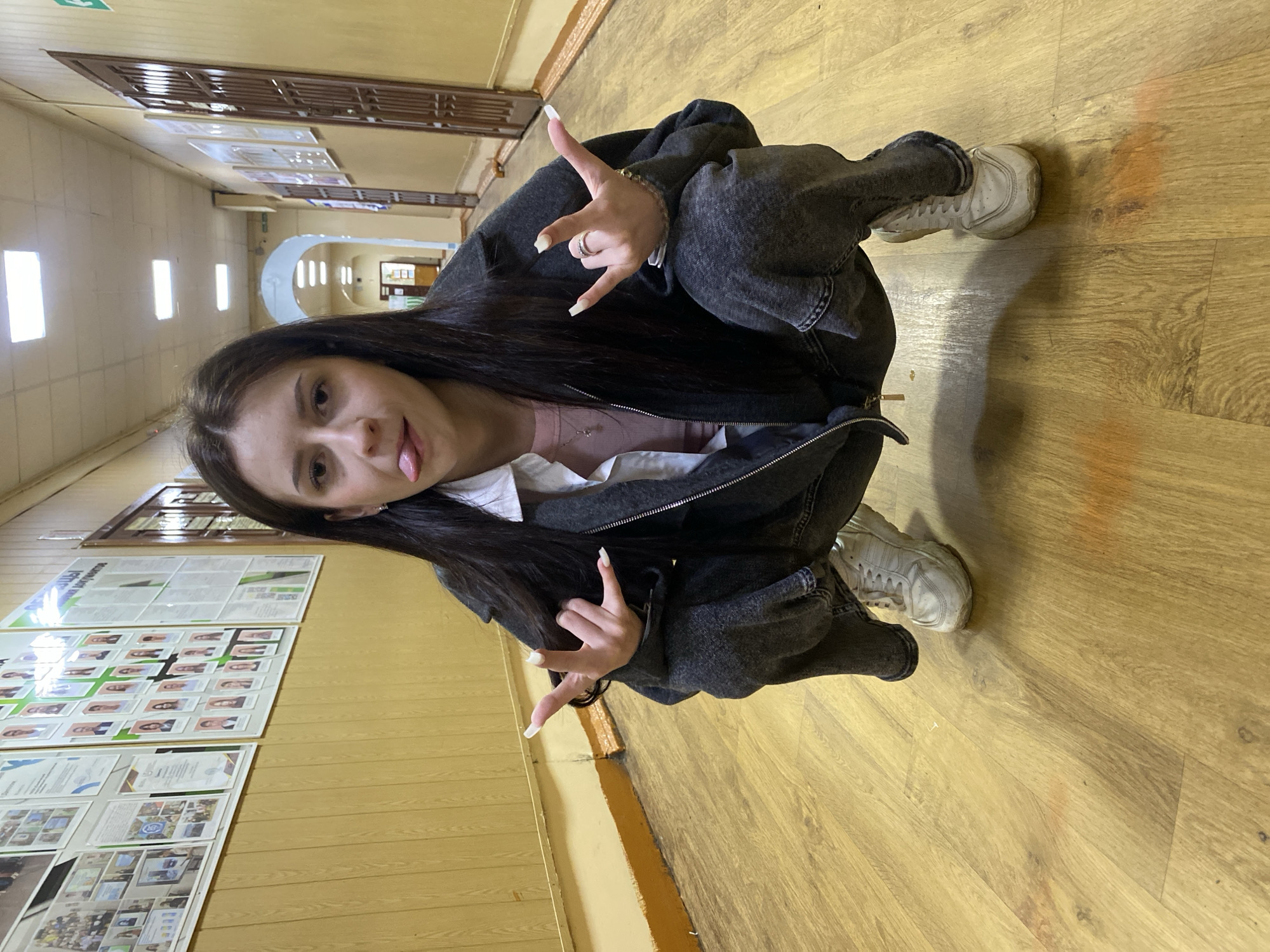 A young woman with long dark hair is captured in a playful pose within what appears to be an indoor hallway. She is squatting or crouching on a light-colored wooden floor, facing the viewer directly. She is making a "rock and roll" or "devil horns" hand gesture with both hands, extending her index and pinky fingers while her thumb, middle, and ring fingers are curled. Her hands feature prominent long white nails. Her facial expression is mischievous, with her tongue slightly sticking out.
She is dressed in a dark, possibly charcoal or washed-out black, zip-up jacket, which is partially open to reveal a light-colored top underneath (a pinkish shirt over a white collar, possibly a layered look). Her lower half is covered by dark pants and she wears white sneakers. A ring is visible on her right hand.
The setting is a well-lit corridor, likely in a school or public building. The walls are a neutral light color, and the floor is made of wooden planks. On the left side of the hallway, there are several display boards or posters mounted on the wall. Some of these boards contain numerous small portrait photographs, possibly of students or staff, arranged in grids. Other boards show documents, certificates, and images, suggesting achievements or information displays. In the background, an arched doorway leads to another section of the hallway. Fluorescent lighting fixtures are visible on the ceiling. The overall impression is one of casual youthfulness in an institutional environment. Visible text on the wall displays is largely unreadable due to distance.