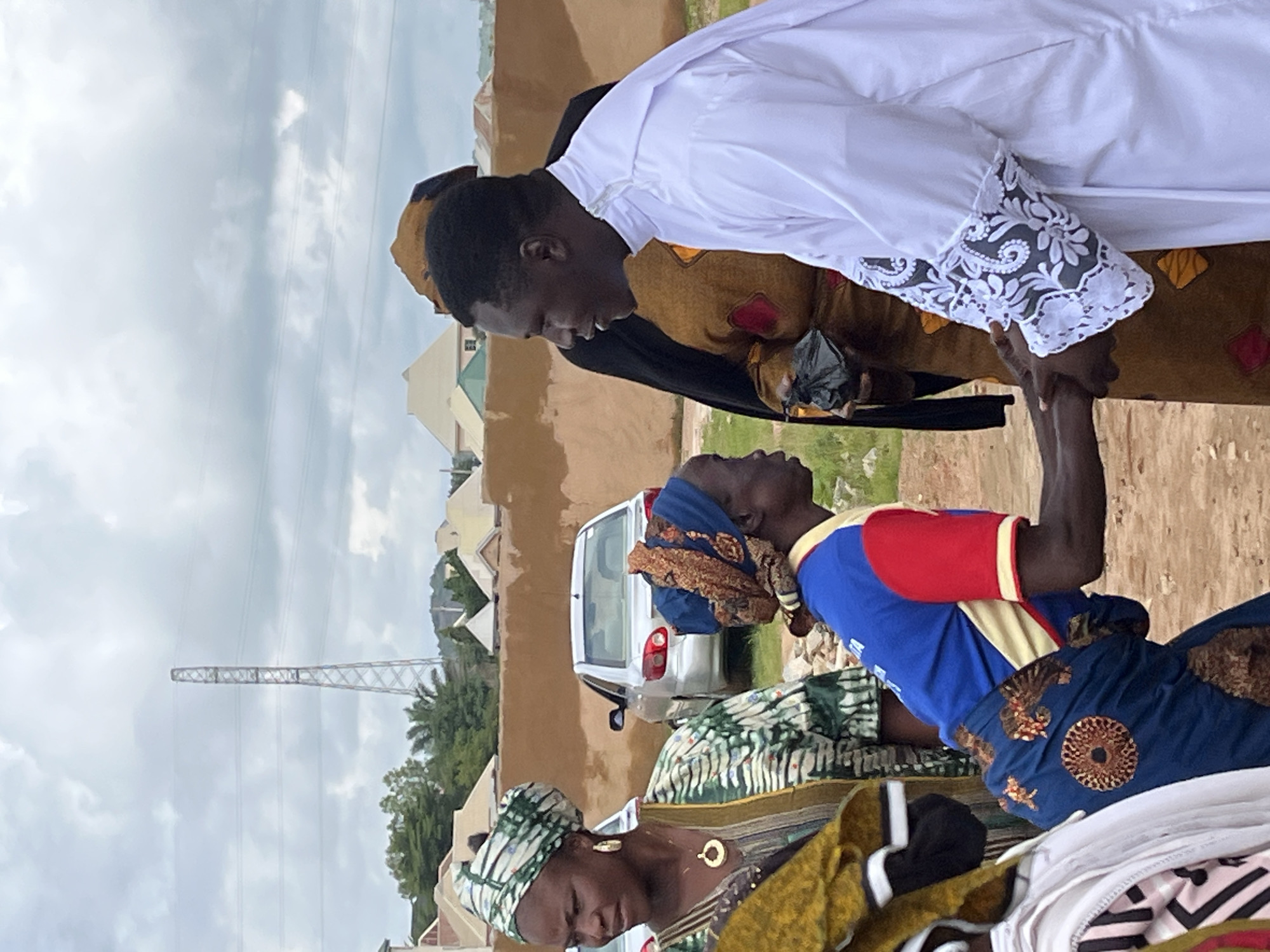 In Jos, Nigeria, a religious ceremony unfolds outdoors under a cloudy sky. A group of people are gathered, with the focal point being an interaction between a man in a white robe and a woman. The man, dressed in a white robe with intricate white lace detailing on the sleeves, is extending his hands. Facing him, a woman is bowing, her hands clasped. She wears a colorful outfit consisting of a blue shirt with red sleeves and a blue skirt adorned with a circular pattern and brown accents. Her head is covered with a patterned blue and orange headscarf. To the left, another woman is visible, wearing a green and white patterned headscarf and a matching patterned garment, her left hand resting on her chest. Behind them, a silver car is parked, and in the background, residential buildings with varying roof colors are scattered across the landscape, with a prominent electricity pylon visible in the distance. The ground appears to be dirt and gravel.