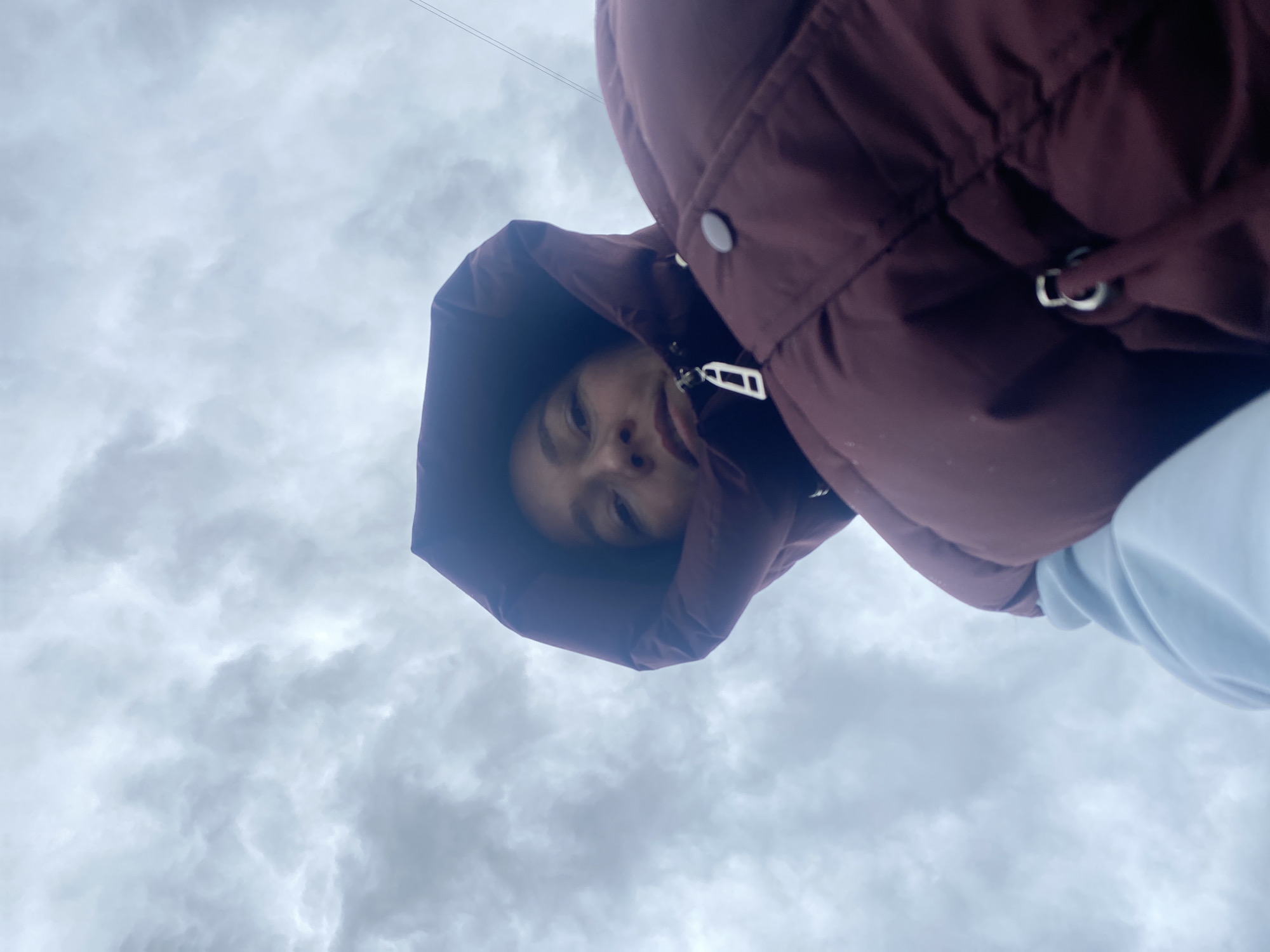 The image shows a person wearing a maroon hooded jacket looking down at the camera. The background is a cloudy sky. There is no information in the image to indicate that it was taken in Chaikovsky, Russia.
