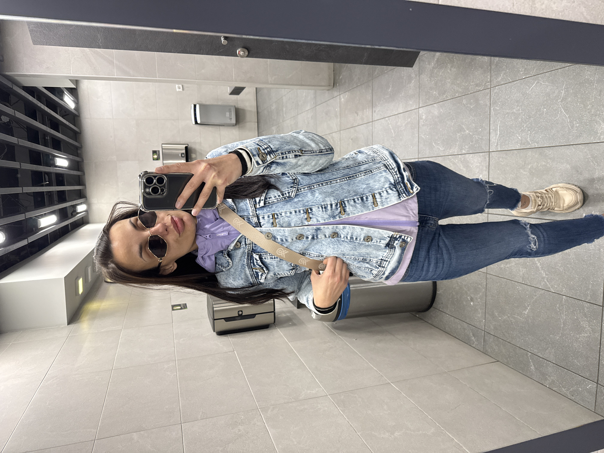 A woman with long dark hair, wearing sunglasses, a light purple hoodie, a light blue acid-wash denim jacket, dark blue jeans with rips at the knees, and light tan sneakers is taking a selfie in what appears to be a public restroom. She is holding a black smartphone in her right hand. She is wearing a crossbody bag with a light tan strap with a pattern. The restroom has light gray tile flooring and walls, with black accents on the ceiling and fixtures. There is a metal trash can next to her on her right. There is a sink and mirror behind her, and a hand dryer and other restroom fixtures on the wall. The location is not identified. No information suggests the city of Sofia, Bulgaria.