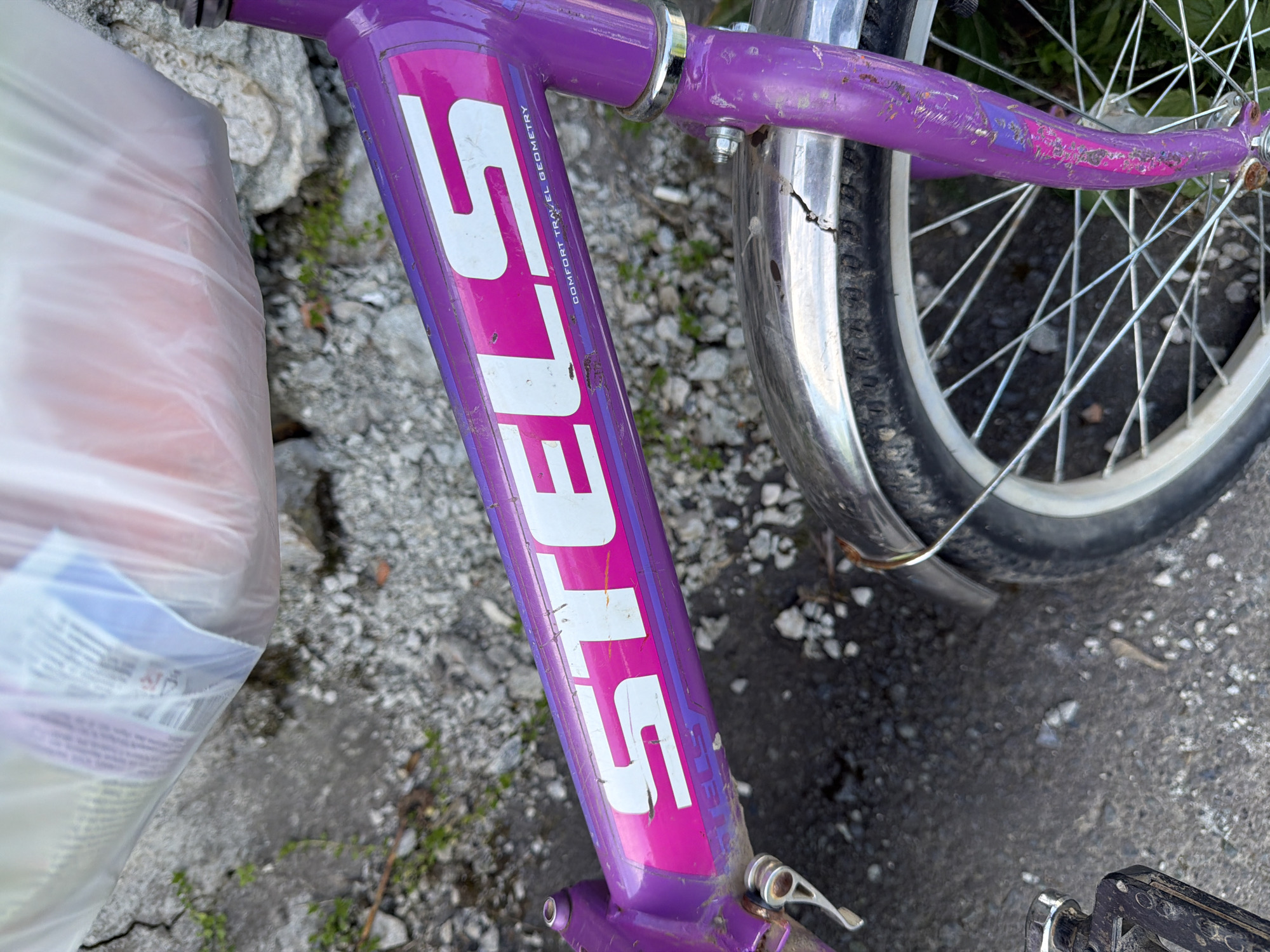 The image captures a close-up, high-angle view of a stationary bicycle frame and parts, situated outdoors on what appears to be a paved or gravelly surface with some weeds.
The primary subject is a purple bicycle frame with prominent pink accents and white text. The paint on the frame shows considerable wear, with numerous scratches and chips revealing the underlying metal or primer. Large white letters spelling "STELS" are visible vertically along the main downtube of the frame. Above "STELS," smaller white text reads "COMFORT TRAVEL GEOMETRY." A silver clamp or ring is visible near the handlebars, which are out of frame. Further down, a silver quick-release lever is attached to the frame.
To the right of the frame, a bicycle wheel is partially visible, showing a silver fender, numerous spokes, and a black tire with a white rim. The wheel appears to be a front wheel. At the bottom right corner, a black bicycle pedal is slightly visible.
On the left side of the frame, a large, translucent white plastic bag rests against the ground, partially obscuring the scene. The bag appears full or partially full, and some blurred contents are vaguely visible through its material.
The ground beneath the bicycle is uneven, composed of a mix of dark grey pavement or dirt, light-colored gravel, and scattered patches of green weeds. The lighting suggests it is daytime, with bright, natural light illuminating the scene. No people are visible, and no specific activity is taking place; the bicycle appears to be parked or leaning.