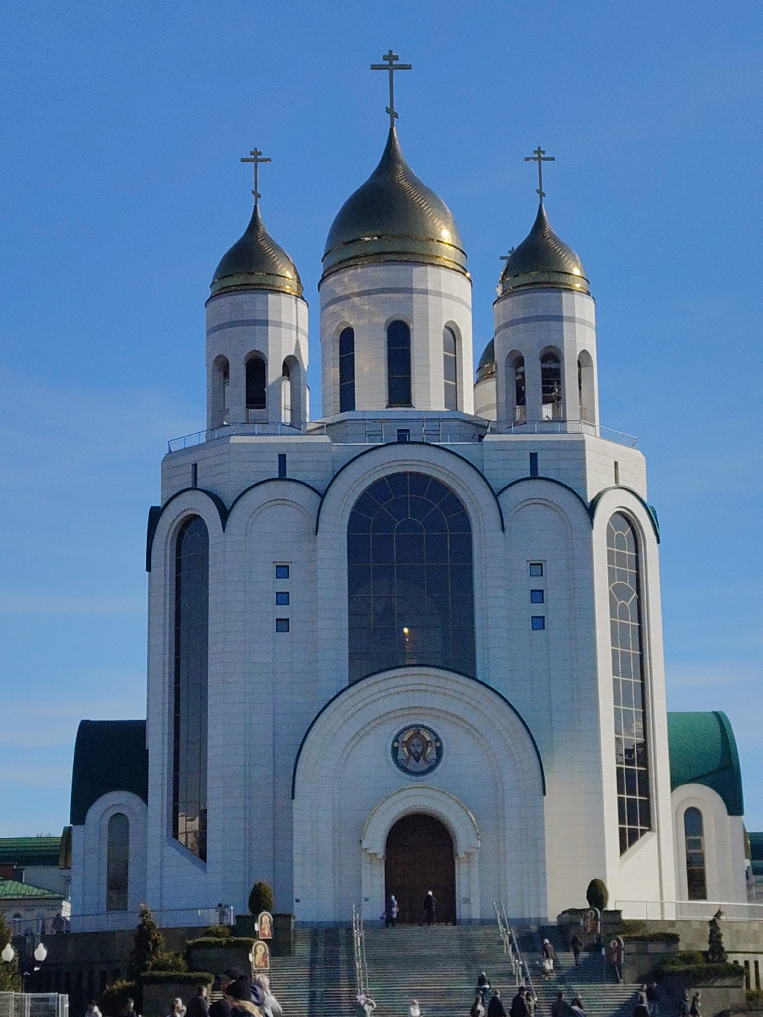 The image depicts the Church on Blood in Yekaterinburg, Russia, under a clear blue sky. The church is a white structure with three prominent golden domes, each topped with a cross. The façade includes large arched windows and a central entrance with a smaller circular image above it. People are seen walking on the stairs leading up to the church. The foreground shows landscaping and some streetlights.