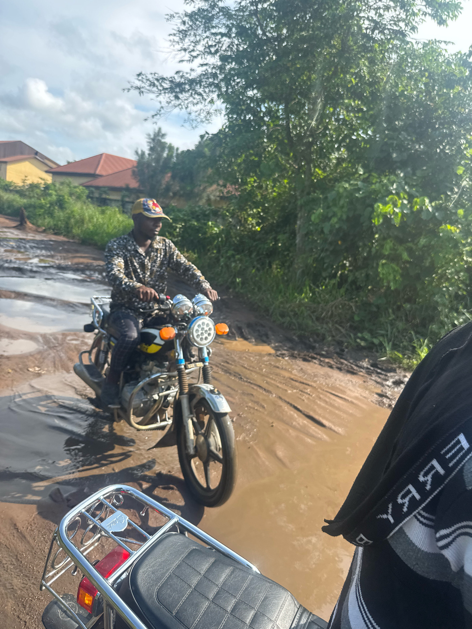In Ihiagwa, Nigeria, a man is riding a motorcycle through a muddy, pothole-filled road. The man is wearing a patterned shirt and a baseball cap. The motorcycle is a modern, street-style bike with a prominent headlight and turn signals. The road appears to be unpaved and in poor condition, with large puddles of water and mud. Lush green vegetation lines the sides of the road, and in the background, there are houses with orange roofs. The sky is partly cloudy. The photo is taken from the perspective of someone on another motorcycle, capturing the front of their vehicle in the foreground.