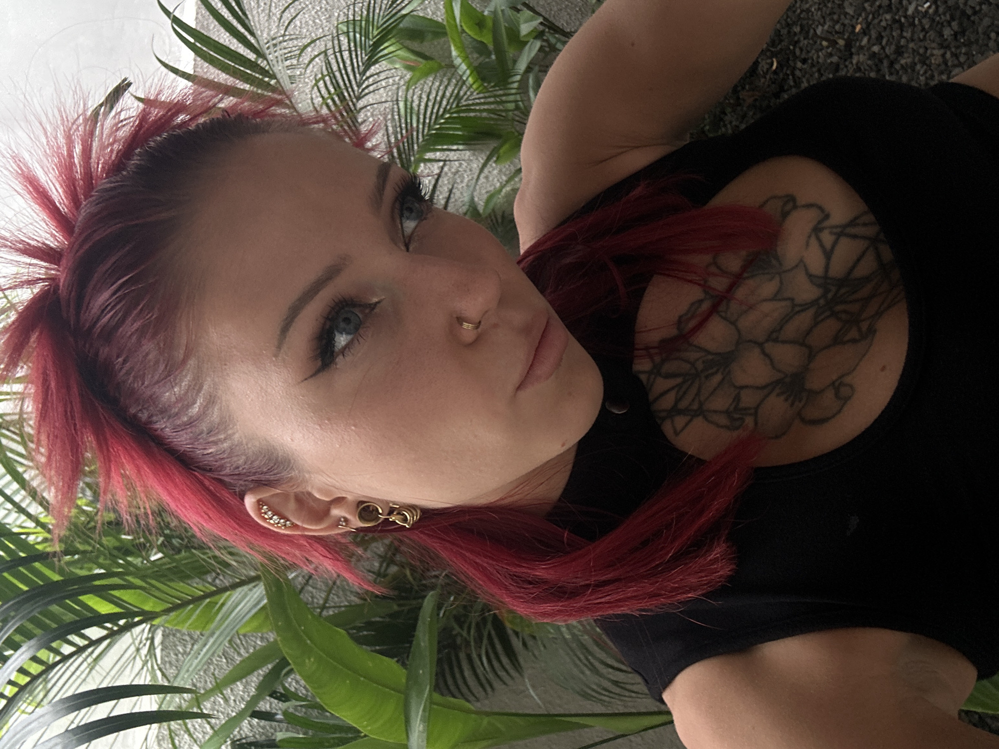 A woman with red hair and multiple ear piercings is seen from a low, upward angle. She is wearing a black top and has a large floral tattoo on her upper chest. Her hair is short on top and longer in the back, with a spiky texture. The background consists of dense green tropical plants, with a surface of dark, gravel-like material visible at the top. The location is Dalung, Indonesia.