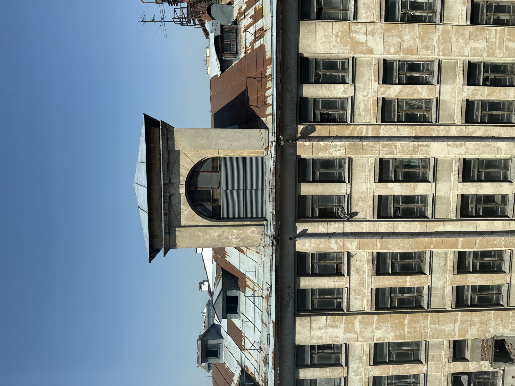 The image shows a building in Moscow, Russia, with a weathered facade and many windows. The building is likely old, judging by its design and condition. There's a prominent architectural feature resembling a lookout or vent on top of the building. Some rusty metal structures are also visible. The sky is clear and blue.