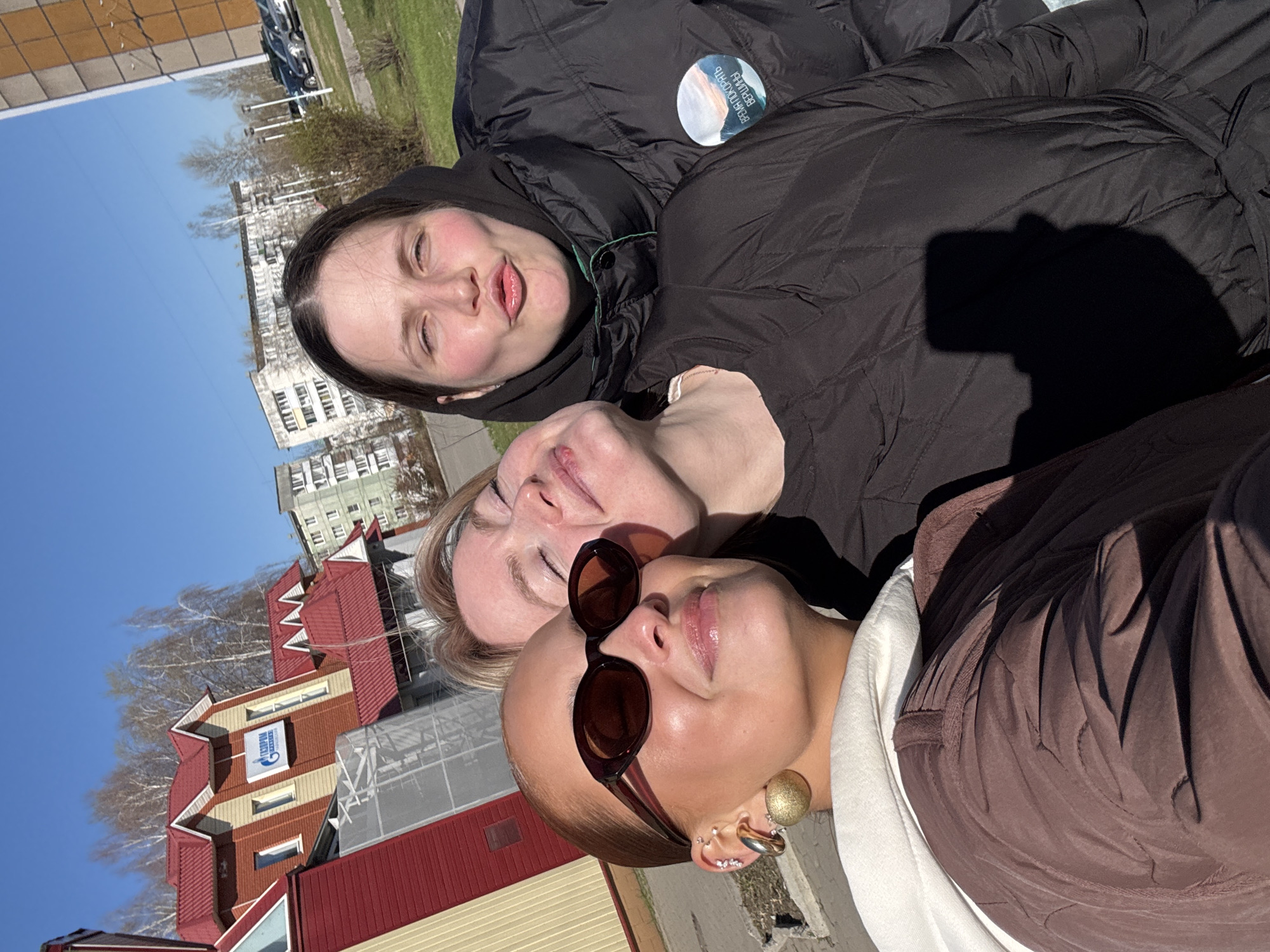 Three women are close together in a selfie outdoors on a sunny day. The woman at the bottom is wearing sunglasses, a brown top, and an earring. The middle woman has blond hair and a light complexion. The woman at the top is wearing a black puffer jacket with a patch. The shadow of a cell phone can be seen near the top right of the image. Buildings are visible in the background, and a sign says "Gazprom," placing the location in Chaikovsky, Russia.