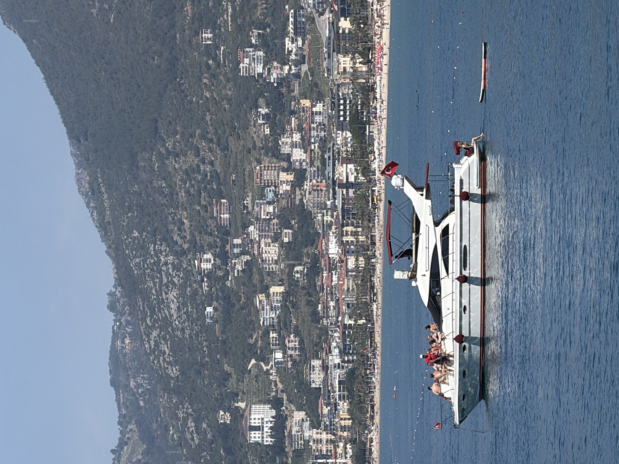 This image depicts a bustling coastal scene in Alanya, Turkey, on a clear, sunny day. In the foreground, a large white motor yacht with red accents is positioned on calm, blue waters. Several individuals are visible on the yacht's deck, relaxing and sunbathing in swimwear. A Turkish flag, featuring a white star and crescent on a red field, proudly flies from the boat's bow. A red and black inflatable object floats near the yacht in the water.
The mid-ground reveals a long, sandy beach, densely populated with numerous sunbathers and beachgoers, along with many beach umbrellas and sun loungers. Behind the beach, a vibrant townscape unfolds, with a multitude of residential and commercial buildings extending along the coastline and climbing the lower slopes of a large, tree-covered mountain that dominates the background. The mountain is lush with greenery, and a few buildings are scattered higher up its slopes.
The weather is bright and sunny, indicative of a pleasant day for outdoor activities. The water is tranquil, with small white buoys marking areas possibly for swimming or navigation. The overall impression is one of leisure and vacation, characteristic of a popular Mediterranean resort. No visible text is discernable apart from the emblem on the Turkish flag.