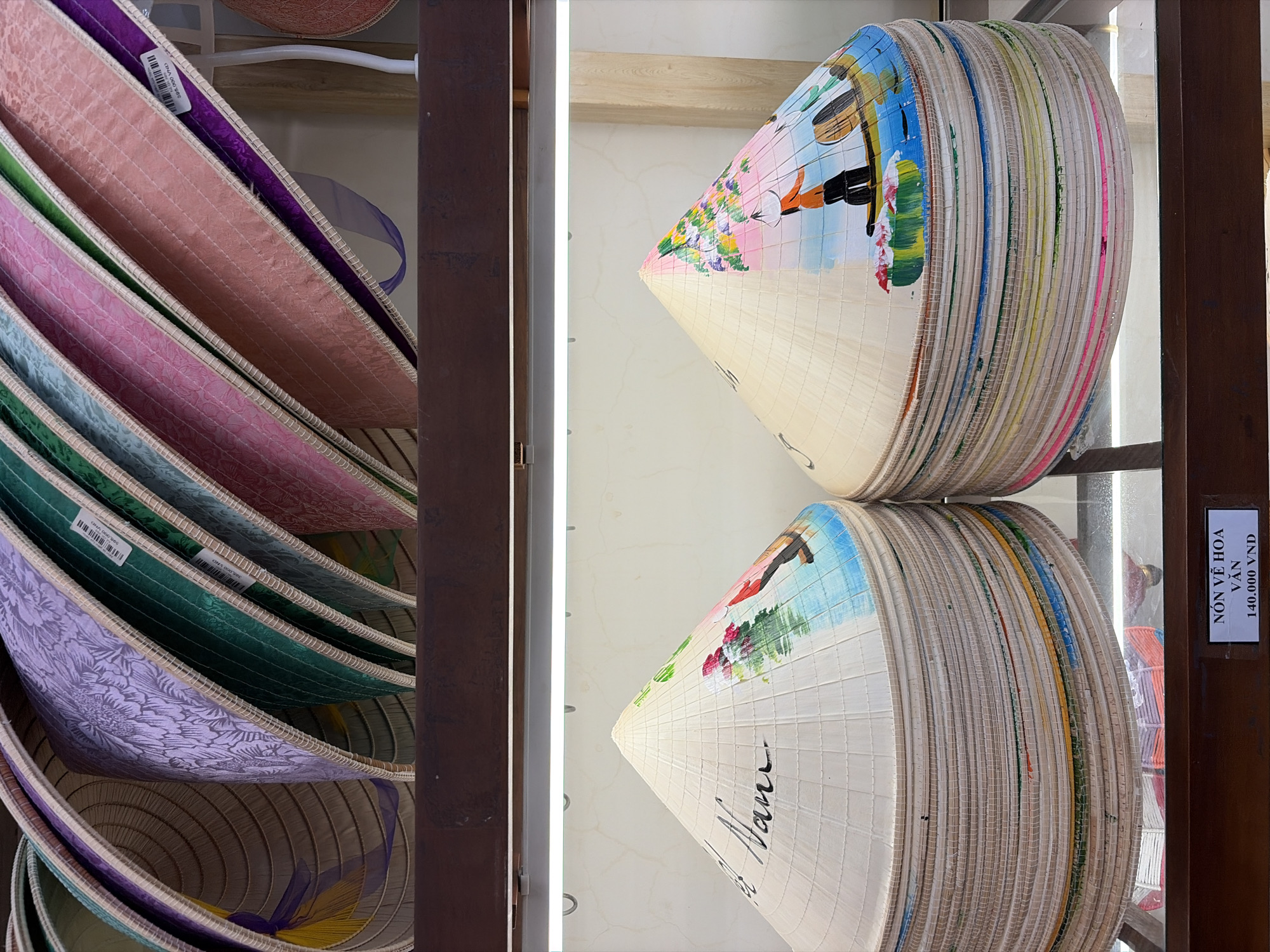 The image shows a display of Vietnamese conical hats, known as "nón lá," in what appears to be a market or shop. The top portion of the image features a close-up of several nón lá with decorative fabric covers in various colors and patterns, including purple with a floral design, green, light blue, and pink. The bottom portion displays a stack of white nón lá that are hand-painted. One hat prominently features the text "Nam" in black calligraphy and painted flowers and a boat scene with figures. The other hat also has a painted scene of a boat with figures. A price tag in the bottom right corner reads "NÓN VẼ HOA VÂN 140.000 VNĐ," indicating these are painted hats priced at 140,000 Vietnamese Dong. The setting suggests an indoor retail environment with artificial lighting. It is not possible to definitively determine the specific location within Da Nang, Vietnam, based solely on the visual information provided.