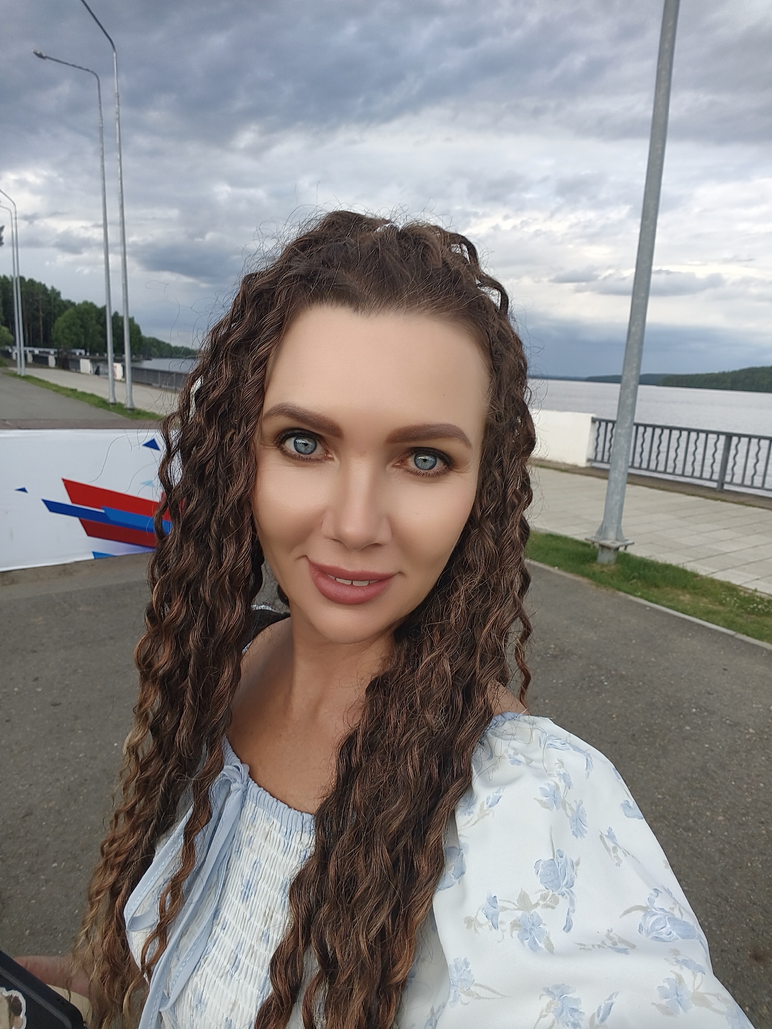 This is a selfie of a woman with long, curly brown hair and blue eyes. She is wearing a light blue and white floral off-the-shoulder top. Her makeup appears to be natural, with a light application of lipstick. She is smiling warmly at the camera.
The background suggests an outdoor setting, possibly a promenade or park by a body of water. A paved walkway is visible behind her, bordered by grass and a metal railing. In the distance, a body of water stretches out, with a tree-lined shore on the far side. The sky is overcast with dramatic, dark clouds, hinting at approaching rain or a stormy day. The time of day appears to be late afternoon or early evening due to the lighting.
To the left of the woman, a white banner with abstract red, white, and blue geometric shapes is partially visible. Several tall, slender light poles are also present in the scene, spaced along the walkway. The overall mood is pleasant and relaxed, despite the cloudy weather.