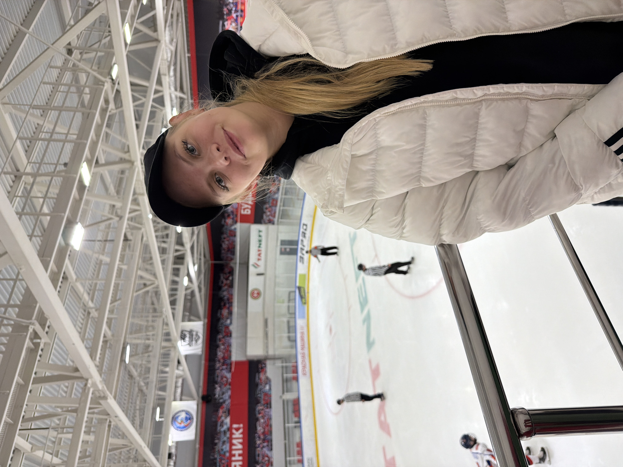 A slightly canted medium shot captures a young, fair-skinned woman with blonde hair and blue eyes, wearing a black hoodie, a white puffy jacket with Adidas stripes, and a black baseball cap. She is smiling slightly at the camera. Behind her is the interior of a hockey arena, specifically the ice rink. There are several referees on the ice. On the walls surrounding the rink are red banners and advertisements. The ceiling of the arena is a complex network of white metal beams and lights. Based on the "TATNEFT ARENA" signage, it is located in Almetyevsk, Russia.