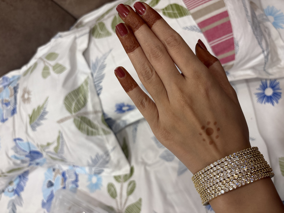 A hand with henna-painted fingertips and a sun-shaped design on the back rests on a bed. The nails are painted a deep red. A wide, multi-strand gold bracelet adorned with sparkling stones encircles the wrist. The bed is covered in a white floral-patterned sheet with blue, green, and gray accents. The background is a blend of the floral sheet and a brown sofa.