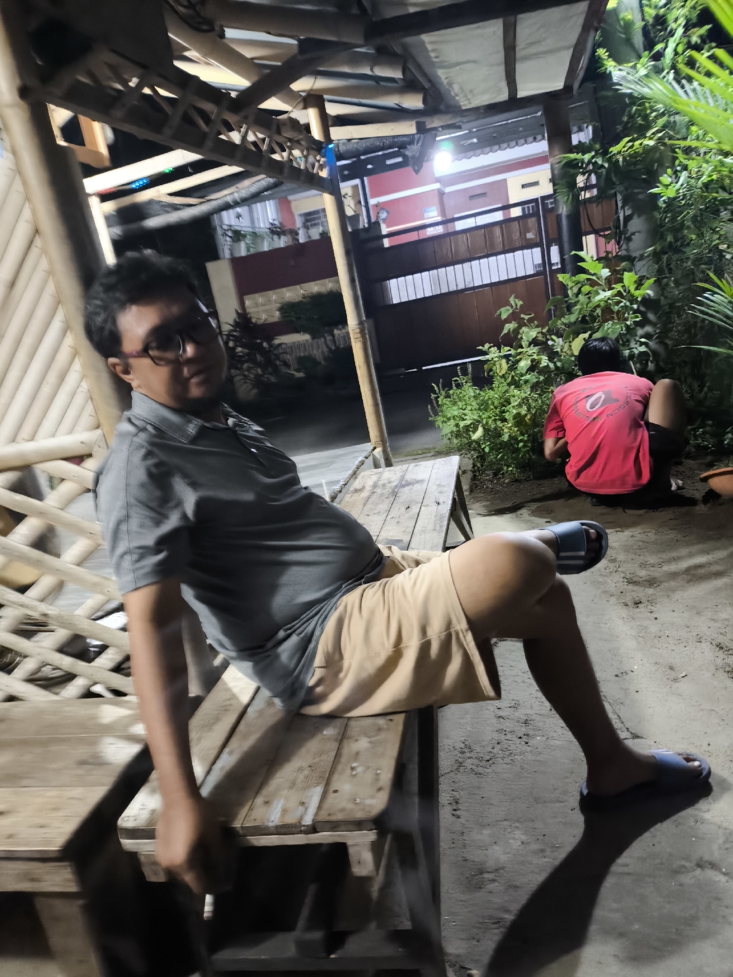 In the city of Cilegon, Indonesia, two individuals are observed in an outdoor, dimly lit setting, possibly a residential backyard or communal area. The foreground features a man of Asian ethnicity, wearing glasses, a grey collared shirt, and light brown shorts, seated on a rustic, wooden bench. His body is turned slightly away from the camera, with his right leg extended and resting on the bench. He appears to be holding a cigarette. To the right of the bench and further back, another person, whose head and upper body are visible, is wearing a red t-shirt and dark shorts. This individual is crouched down, facing away from the camera, tending to or observing a cluster of green plants. The environment is characterized by sandy or dirt ground, and a bamboo-constructed shelter with a wooden roof is visible in the background, along with some foliage and a gated entrance to a building. The lighting suggests it is evening or nighttime.