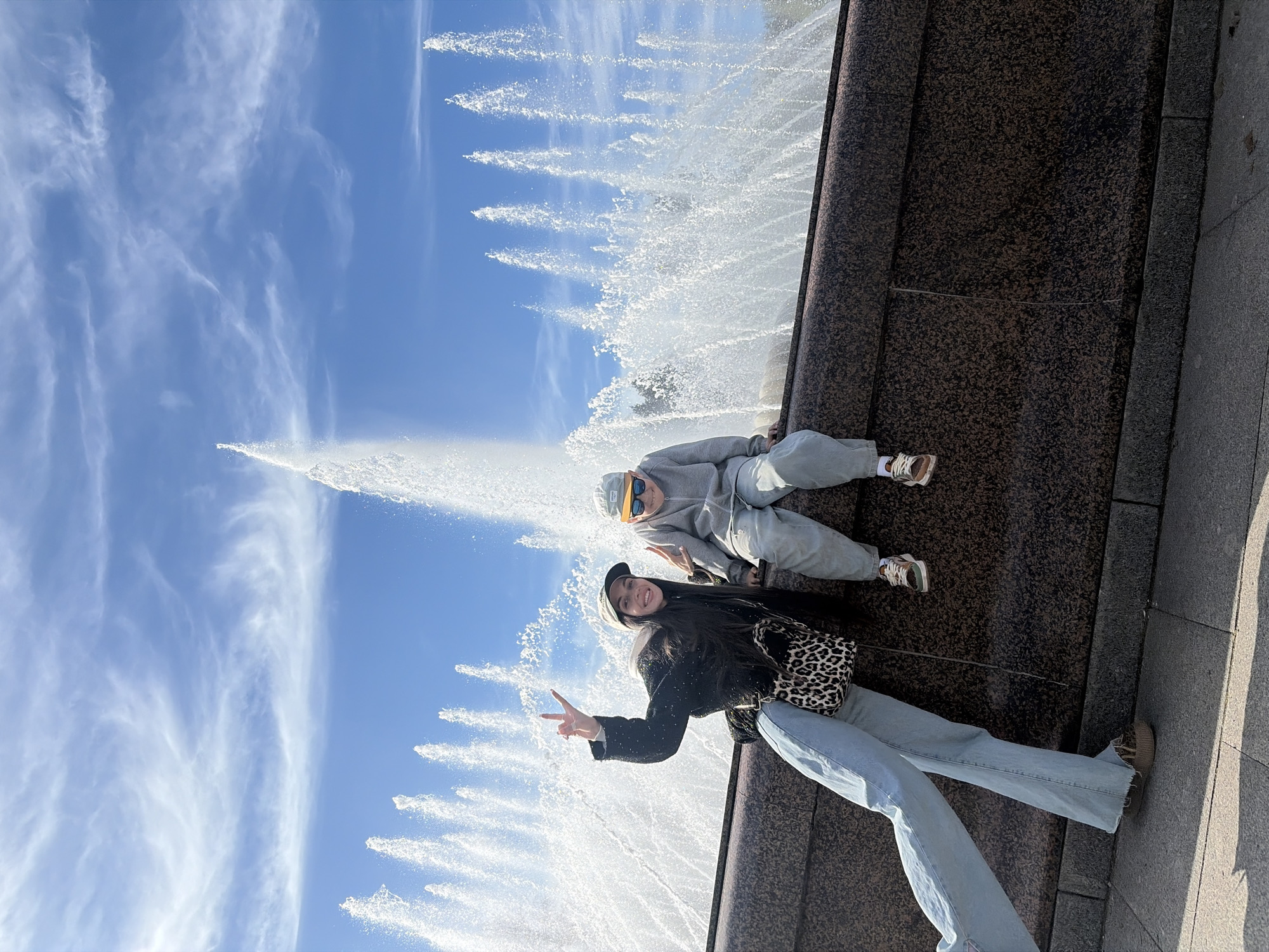 This outdoor photograph, correctly oriented, captures two young individuals posing cheerfully beside a large fountain in Saint Petersburg, Russia, under a bright blue sky.
The scene is dominated by a majestic fountain with numerous powerful water jets creating a dynamic backdrop against a clear sky with sparse, wispy white clouds. On the right side of the frame, a dark, mottled granite-like stone wall forms the edge of the fountain, extending from the bottom to the top of the image.
In the foreground, a person stands facing the camera, smiling and making a peace sign with their right hand. They are dressed in light blue wide-leg jeans, a dark textured jacket, and a light-colored baseball cap. A distinctive leopard print crossbody bag hangs across their body. Slightly behind and to their right, a second person is seated comfortably on the stone edge of the fountain, also looking towards the camera. This individual is wearing a grey hooded tracksuit (hoodie and sweatpants), a light-colored cap, and sunglasses. Their sneakers are visible resting on the stone surface.
The image conveys a casual and happy moment, likely captured during the day, given the clear sky and bright illumination that highlights the splashing water and the details of the subjects' clothing and the stone. No visible text is present in the media file.