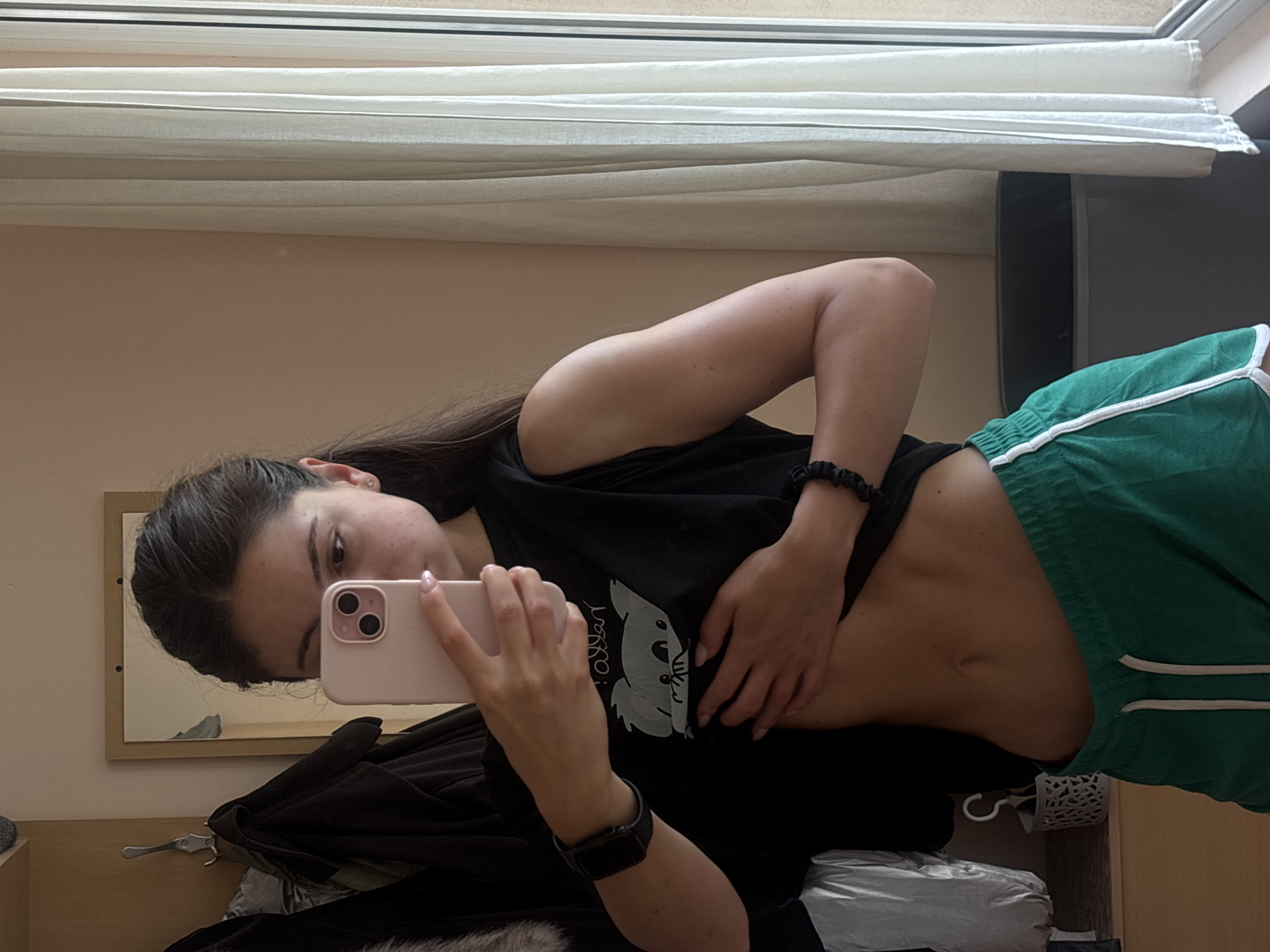 The media file is a vertical, selfie-style photograph taken indoors. The main subject is a young woman with long, dark hair. She is wearing a black t-shirt with a graphic of a koala on it and green shorts with white trim. The woman is holding a pink smartphone in her right hand, seemingly taking the photo. Her left hand is resting on her stomach, exposing her midriff.
The setting appears to be a bedroom or dressing area. In the background, there is a window covered by white curtains. A mirror with a wooden frame is visible to the left, reflecting part of the room and the woman's head and shoulder. There are clothes draped over what appears to be a hanger or rack.
The lighting suggests it is daytime. The woman's expression is neutral, as she focuses on taking the selfie. The image is taken from a slightly low angle, emphasizing her pose. There is no visible text in the image, except for the graphic on her t-shirt, which is stylized and not clearly legible. The overall impression is casual and personal.