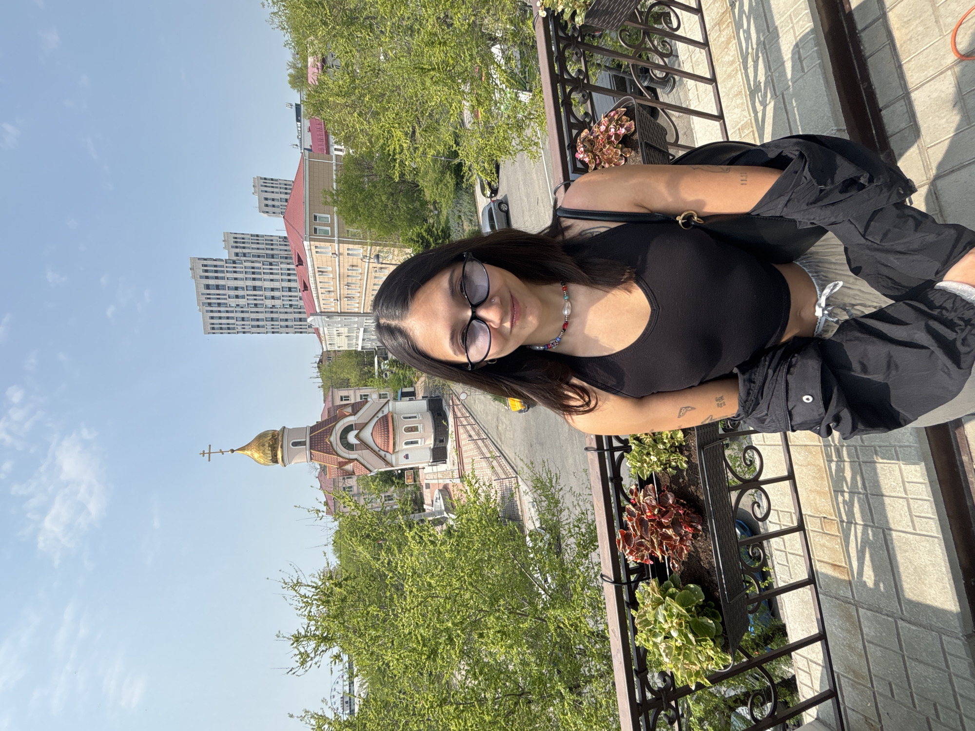 A woman with dark hair and sunglasses stands on a balcony. She is wearing a black top and dark pants, with a purse slung over her shoulder. Balcony flower boxes contain mixed green and red plants. The backdrop reveals a city view including a church with a golden dome and taller buildings. This scene is likely located in Vladivostok, Russia based on the architectural style of the church.