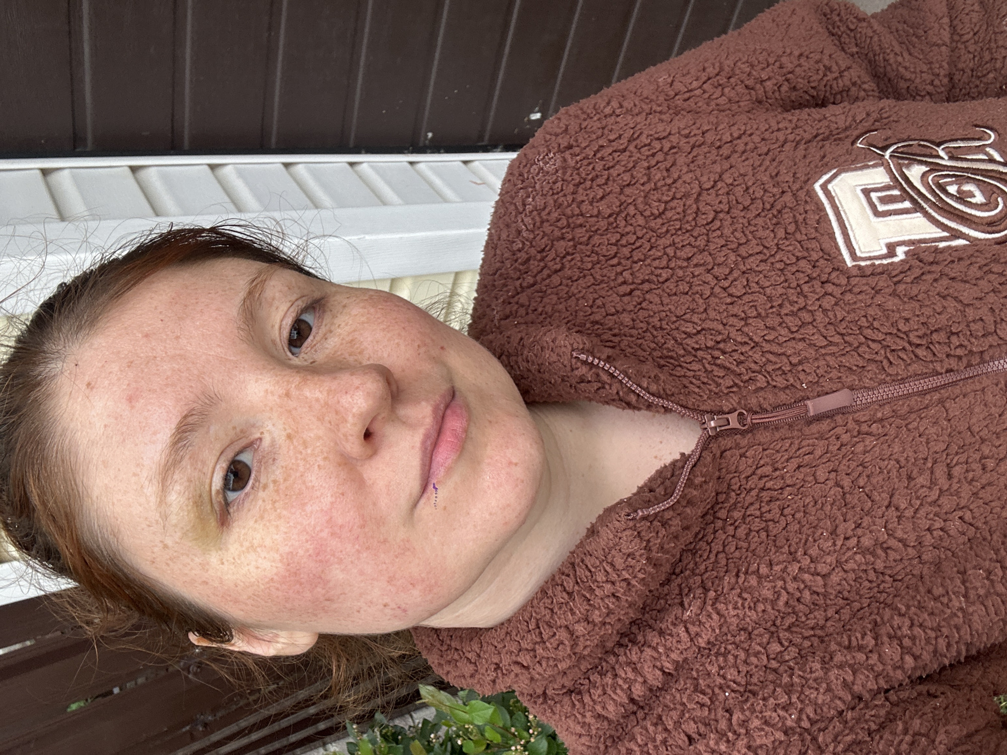 A woman with fair skin, freckles, and reddish-brown hair is wearing a brown, fuzzy zip-up jacket with a white patch on the upper right shoulder. The patch contains a design. The woman has a piercing on her lower lip, and a slight discoloration under her left eye. The background appears to be an outdoor setting with off-white and brown paneling above her, and a bench or ledge below, with green plants visible at the very bottom. The location is unspecified, and there's no clear indication that it is in Nizhny Novgorod, Russia. The image is slightly tilted.