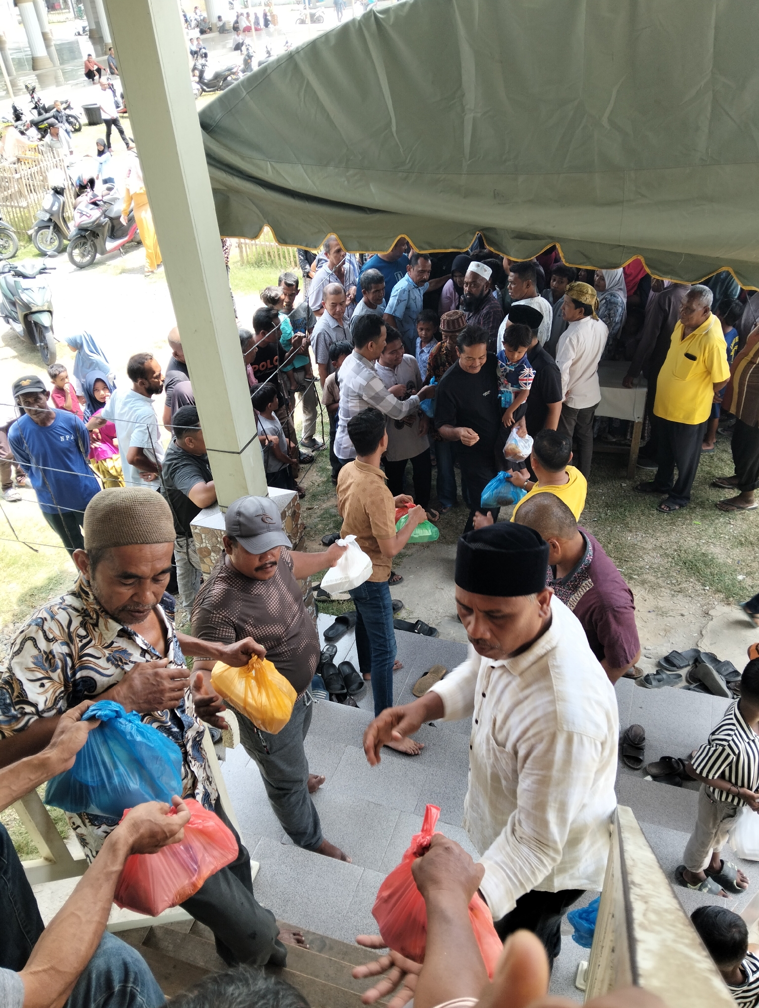 The image depicts a crowded scene, possibly a religious gathering or distribution event, in Grong-Grong, Indonesia. A large group of people, predominantly men and some children, are gathered outdoors under a large, olive-green tent. Many individuals are wearing traditional Islamic attire, such as peci caps and long-sleeved shirts, while others are in casual clothing.
Several people are holding plastic bags, some filled with food or goods, suggesting a distribution or charitable event. In the foreground, a man in a patterned shirt is holding a blue plastic bag, and another man in a white shirt and black peci is reaching for a red plastic bag. Another man in a brown patterned shirt is holding a yellow plastic bag. The ground is a mixture of concrete steps and dirt, with scattered footwear.
In the background, motorcycles are visible, indicating a mode of transportation common in the area. A wooden fence and a white pillar also frame the scene. The lighting suggests it is daytime, with bright sunlight casting shadows. The overall impression is one of community and shared activity, likely centered around a religious or cultural observance.