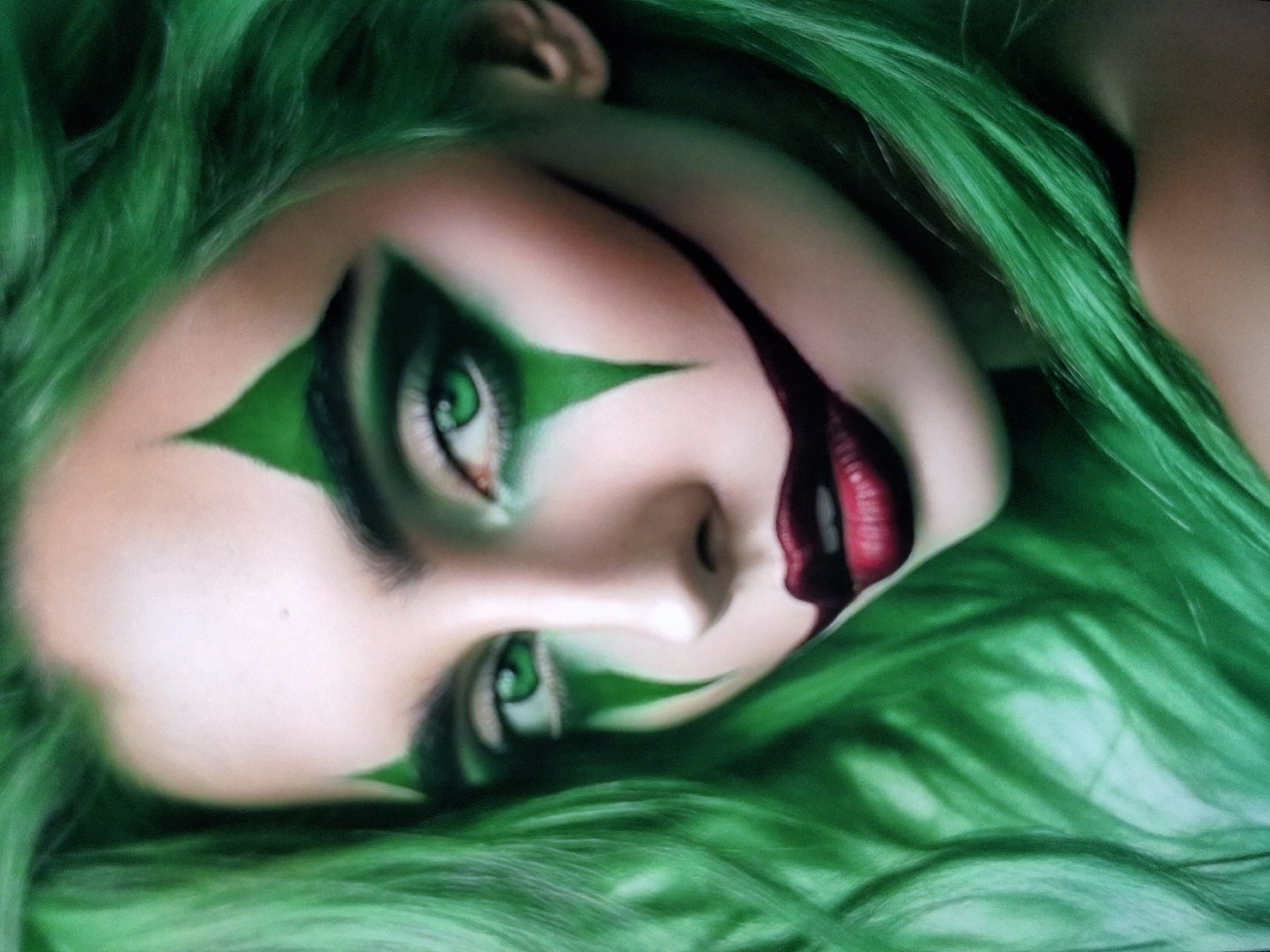 An extreme close-up, head-and-shoulders portrait of a young woman with bright green hair and striking green eyes, wearing clown-like makeup. Her face is adorned with pointed green markings above her eyes and dark red lipstick that extends into a stylized smile. The lighting is soft and diffused, highlighting the contours of her face and the texture of her hair. The background is blurred and abstract, with hints of the same green as her hair. There is no discernible city or environmental context provided.