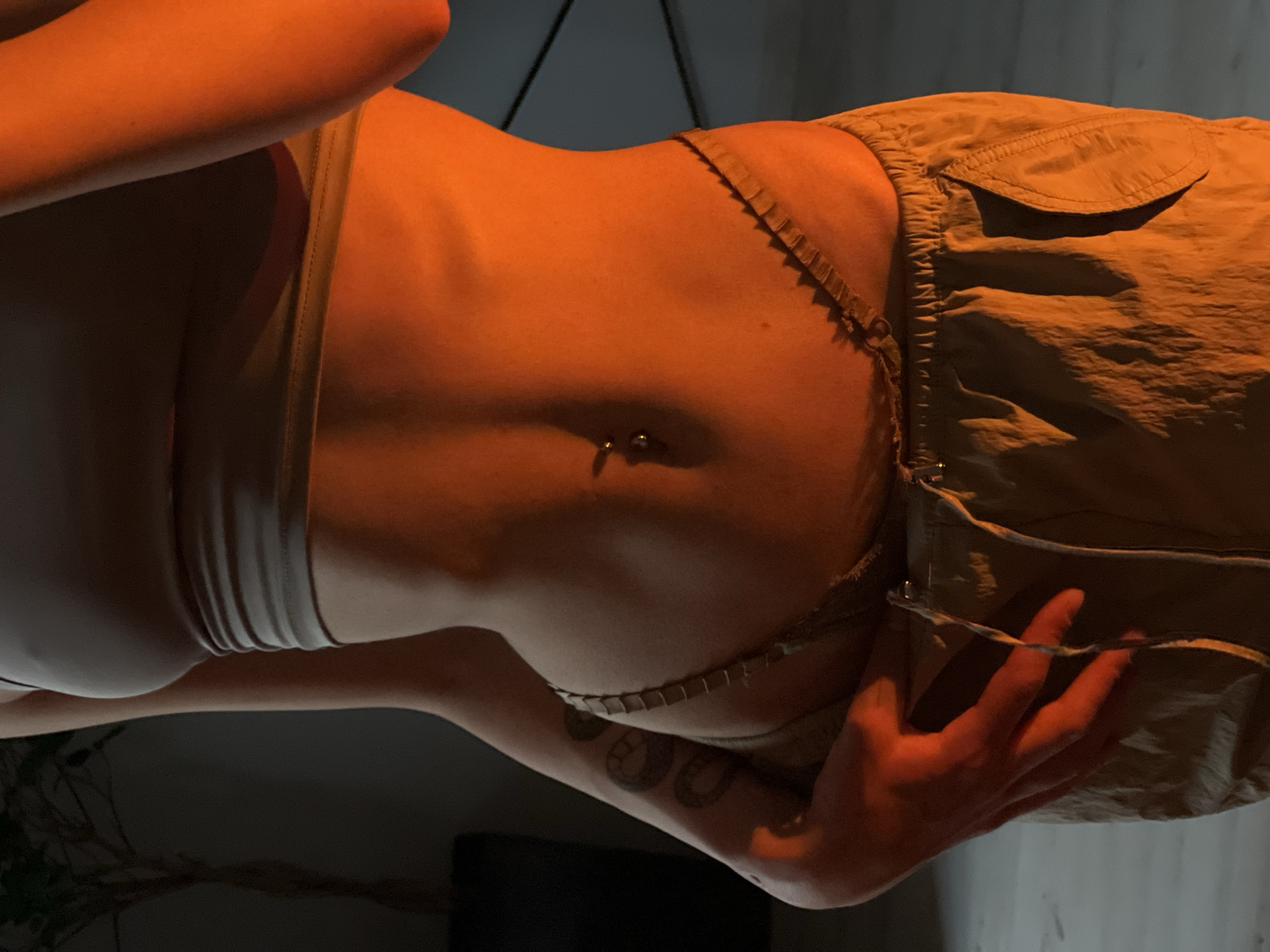 The image is a medium shot of a person's torso, illuminated by warm orange light. The person is wearing a light-colored crop top and beige pants. A navel piercing is visible, as is a tattoo on the left arm. The background is dark with a blurred plant to the left and a hint of a wall or structure to the right. There is no indication of the location being Ternopil, Ukraine.