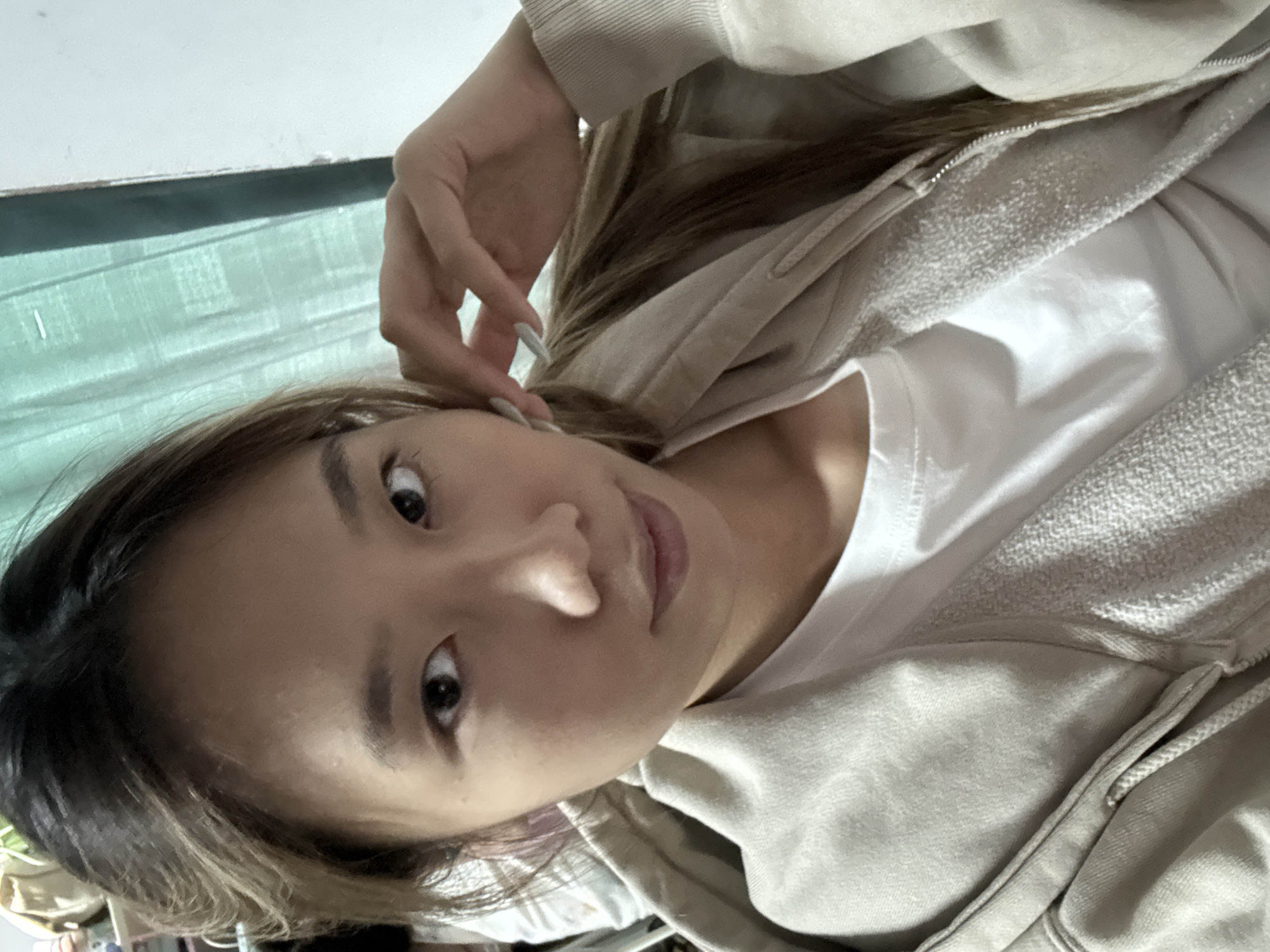 A person with light brown hair and skin is oriented at an angle, facing the camera. The person is wearing a white T-shirt and a light beige hooded sweatshirt. The left hand of the person is near their left ear. The background includes a textured teal surface. The location cannot be determined to be in the city of Nan'an District, China, from the image alone.