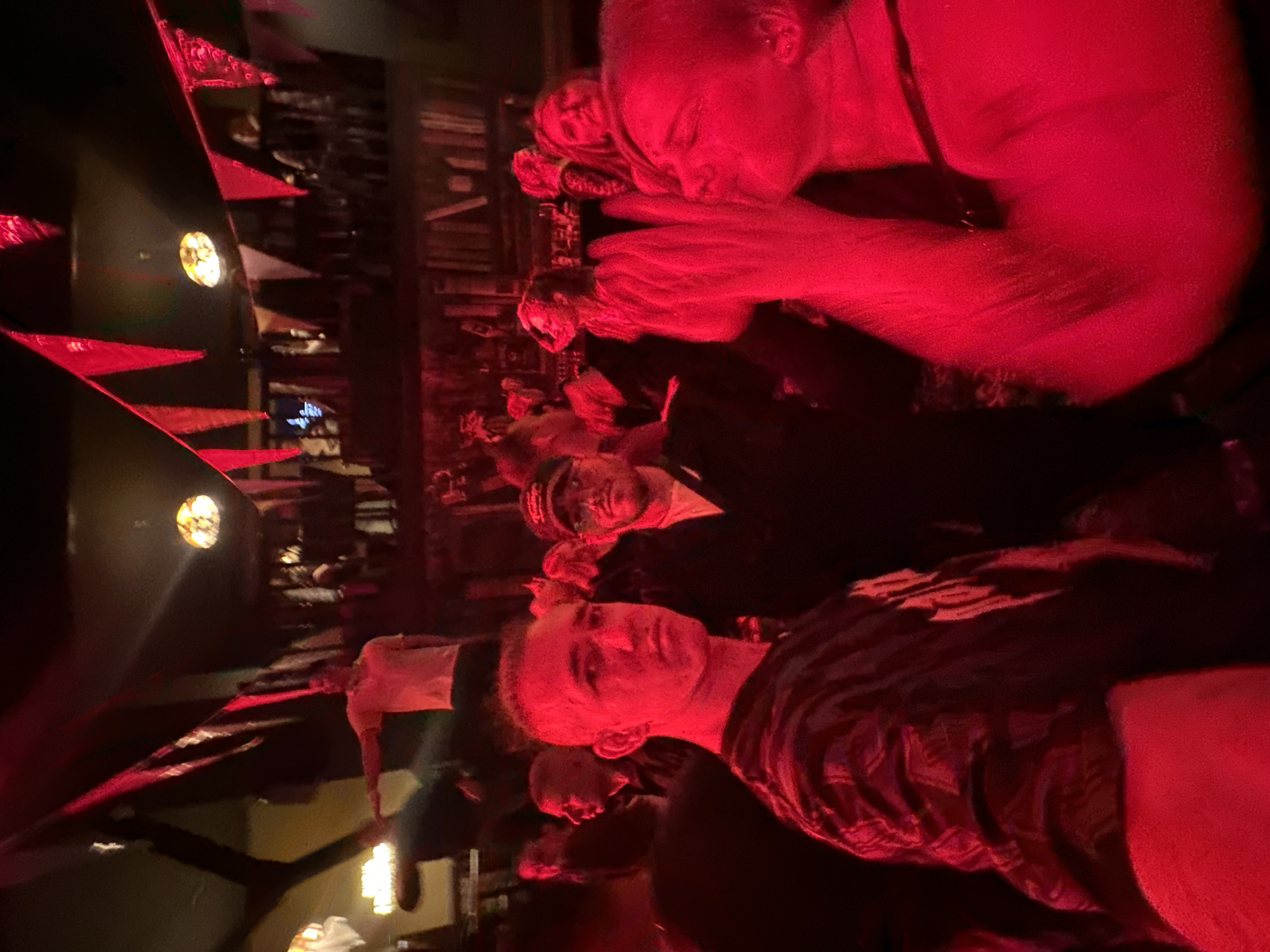 A concert venue in Saratov, Russia is lit with red lighting, illuminating a crowd. Pennants are hung around the interior. A man in the foreground points with his left hand. The crowd is diverse and active. A man in the top right corner holds his hands up to his face.