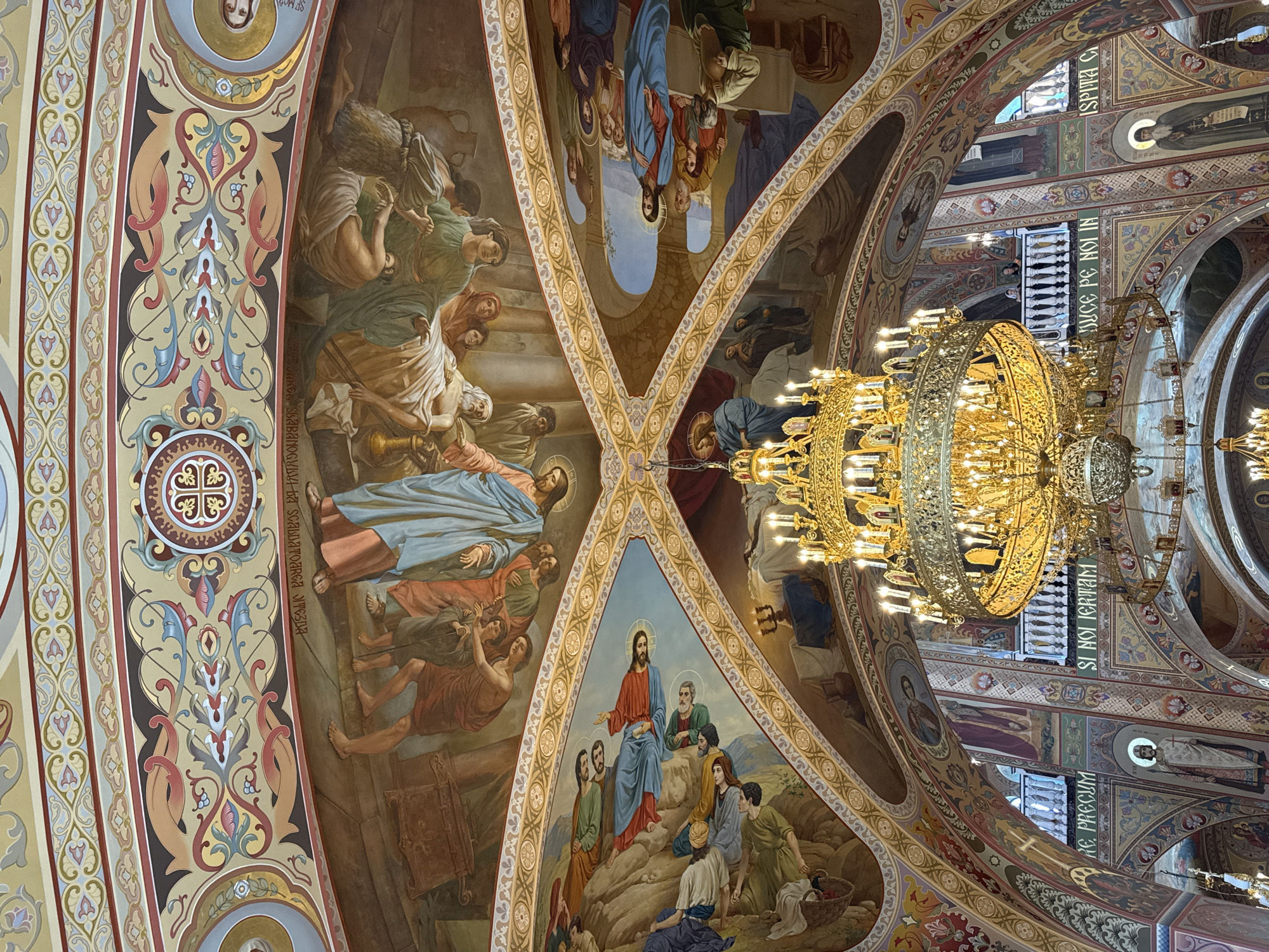 A grand, ornate chandelier hangs from the elaborately painted ceiling of an Orthodox church. The ceiling features frescoes depicting biblical scenes, with figures in flowing robes arranged in dramatic compositions. Swirling patterns and decorative borders in gold, red, blue, and earth tones frame the artwork. The church walls are also adorned with religious imagery and inscriptions in Cyrillic script. Sunlight streams through unseen windows, illuminating the golden structure of the chandelier and casting a warm glow on the vibrant frescoes.