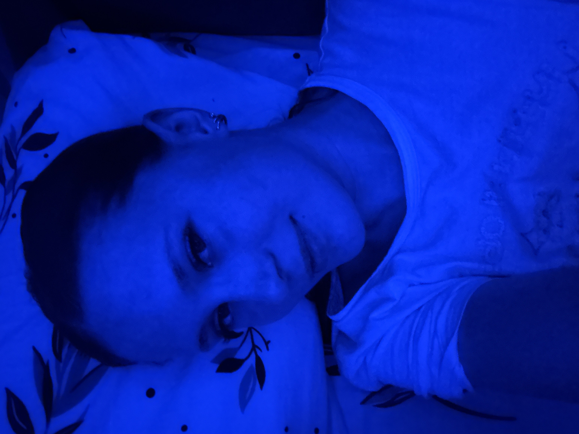 A woman lies on a pillow and bedsheet printed with leaves and dots, under a blue light. No evidence suggests a location in Chaikovsky, Russia.
