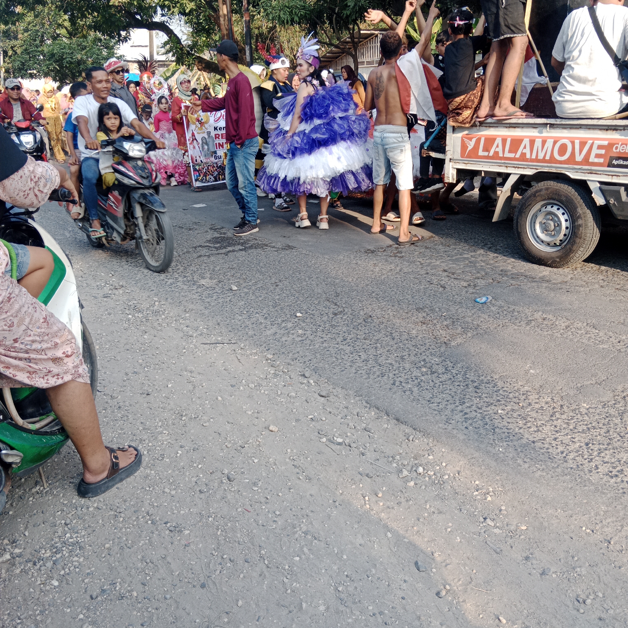 In the city of Tegalrejo, Indonesia, a parade is in progress on a paved street. A group of people, some in festive attire, are participating in the event. A man and a young girl are riding on a motorcycle, with the man in a white t-shirt and the girl in a yellow top. Behind them, a person in a vibrant blue and white ruffled costume is posing. Several other individuals are visible, including those in the background and a person standing on the back of a truck. The truck has "LALAMOVE" written in orange on its side. The scene is framed by trees and a wooden structure. The ground is a mix of asphalt and gravel, with scattered stones. The lighting suggests it is daytime, possibly late afternoon due to the shadows.