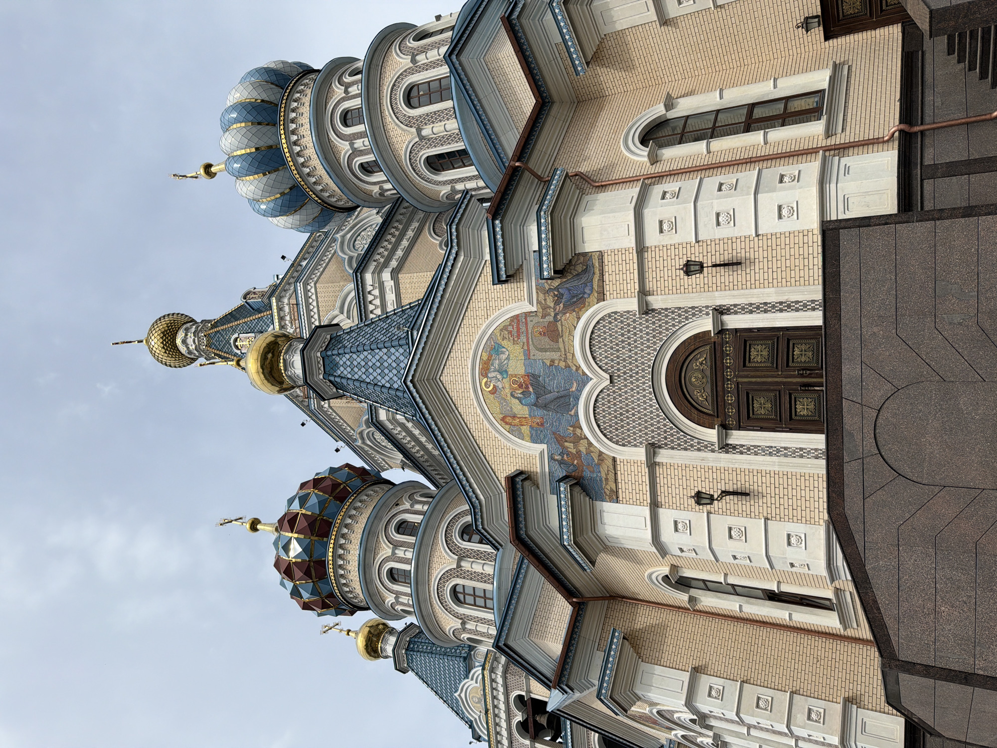 The image captures the exterior of the Alexander Nevsky Cathedral, an Orthodox Christian church located in Izhevsk, Russia. The architectural style is elaborate, featuring a facade of light brown brick adorned with white decorative elements. The building features multiple domes with varied designs. Some domes are blue and white, while others are red and blue, all topped with golden crosses. Above the main entrance, a mosaic depicting religious figures is visible. A copper-colored rain gutter is affixed to the side of the building. The cathedral is bordered by dark granite stone, which appears to be part of the ground or a set of stairs leading to the building. The sky is overcast.
