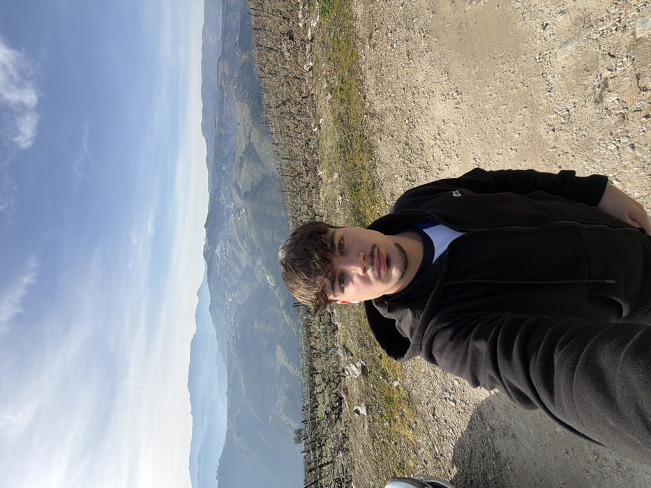 A young man in a black jacket takes a selfie against a scenic backdrop. He stands on a gravel path, adjacent to a steep slope with terraced vineyards. Distant mountains fade into a light blue sky. The sunlight illuminates the path and the man's face, creating shadows that accentuate the rugged landscape.