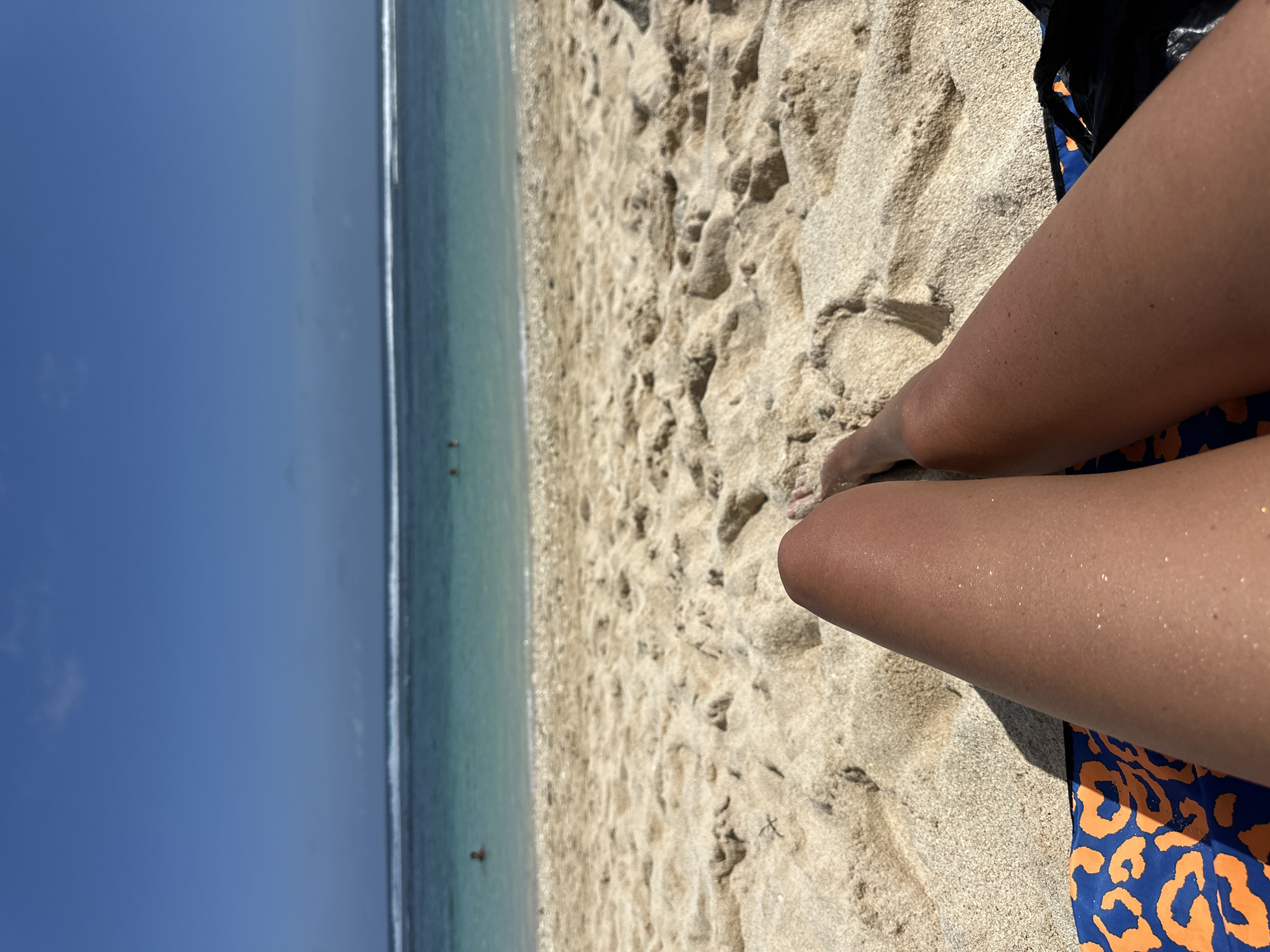 In Kuta Selatan, Indonesia, a person's tanned legs are visible, lying on a beach with sandy ground. The legs are positioned diagonally across the lower part of the frame, with one leg bent at the knee. A blue and orange patterned towel is partially visible underneath the legs. The sand is light brown and textured, with ripples and shadows. In the background, the ocean is a clear turquoise color, with gentle waves breaking near the shore. Several people can be seen swimming or wading in the water. The sky above is a clear, bright blue.