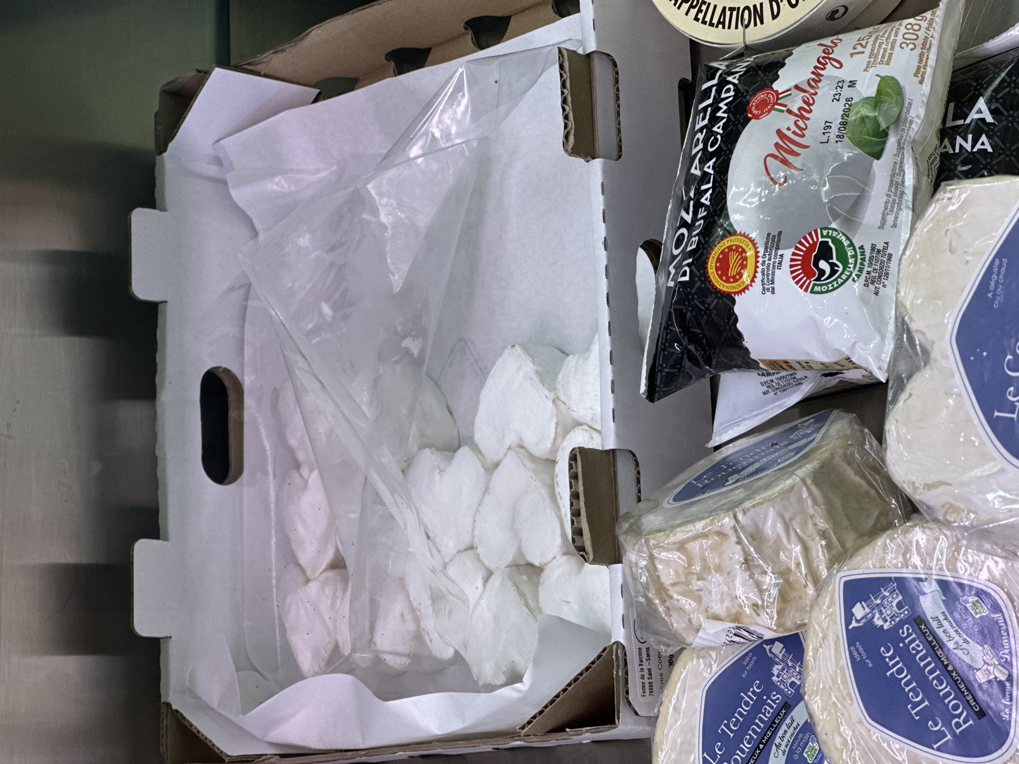 In a retail setting, a cardboard box contains a white paper lining and several heart-shaped cheeses covered in plastic wrap. To the right, a bag of Michelangelo mozzarella di bufala Campana is visible, along with other packaged cheeses. The environment appears to be a cooler or display case, with brushed metal in the background.