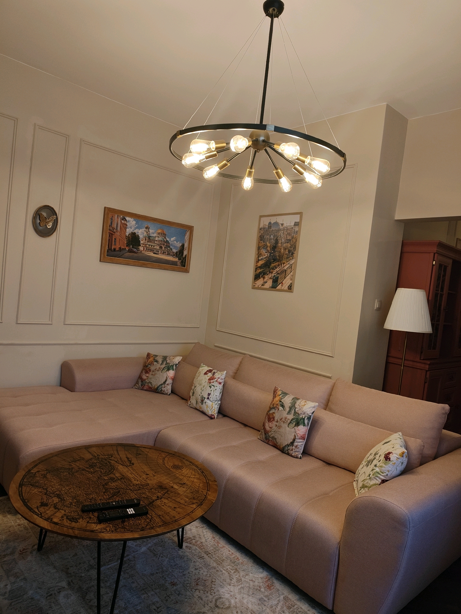 This is an indoor, eye-level, medium shot of a living room in an unknown location. The room is decorated in warm, neutral tones with a pink sectional sofa as the focal point. The sofa is adorned with several decorative pillows featuring floral patterns. In front of the sofa is a round wooden coffee table with a world map etched into its surface. Two black remote controls rest on the table. Behind the sofa, a tall floor lamp with a white shade stands next to a red, glass-fronted cabinet. The walls are adorned with two framed artworks depicting European city scenes. A modern, multi-bulb chandelier hangs from the ceiling, casting a warm glow over the room. The overall impression is one of a comfortable and stylish living space.