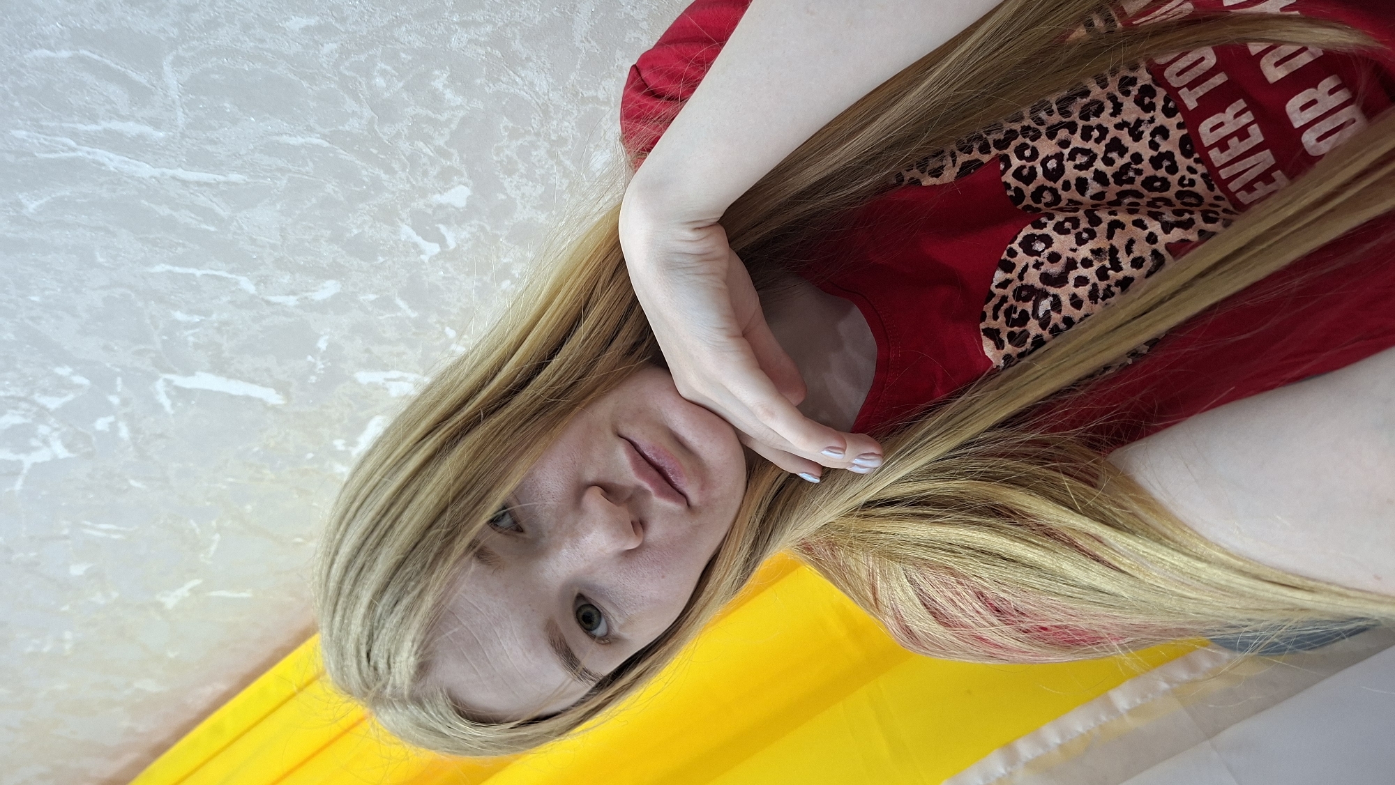 A woman with long, blonde hair is resting her hand on her face. She is wearing a red shirt with a leopard-print heart. A yellow flag is visible at the bottom of the image. The background is a light-colored wall. No information on the city is available.