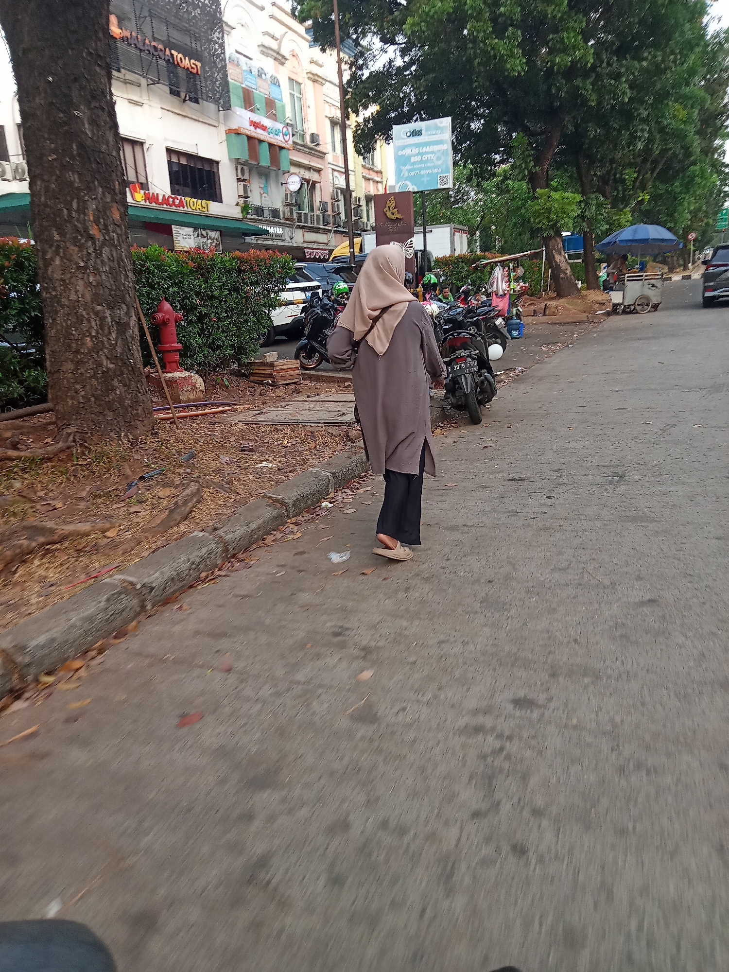 The image captures a street scene in South Tangerang, Indonesia. In the foreground, a woman wearing a light brown hijab and a long, grey tunic with dark pants walks away from the camera on a paved sidewalk. She is wearing light brown slip-on shoes. To her left, a large tree trunk is visible. Behind the woman, several motorcycles are parked along the side of the road. Further back, buildings with signage are visible, including "MALACCA TOAST." A red fire hydrant stands near the curb. In the background, a blue umbrella shades a small vendor cart. The street appears to be lined with trees and other vegetation. The overall impression is that of a typical urban environment with pedestrian and vehicular traffic.