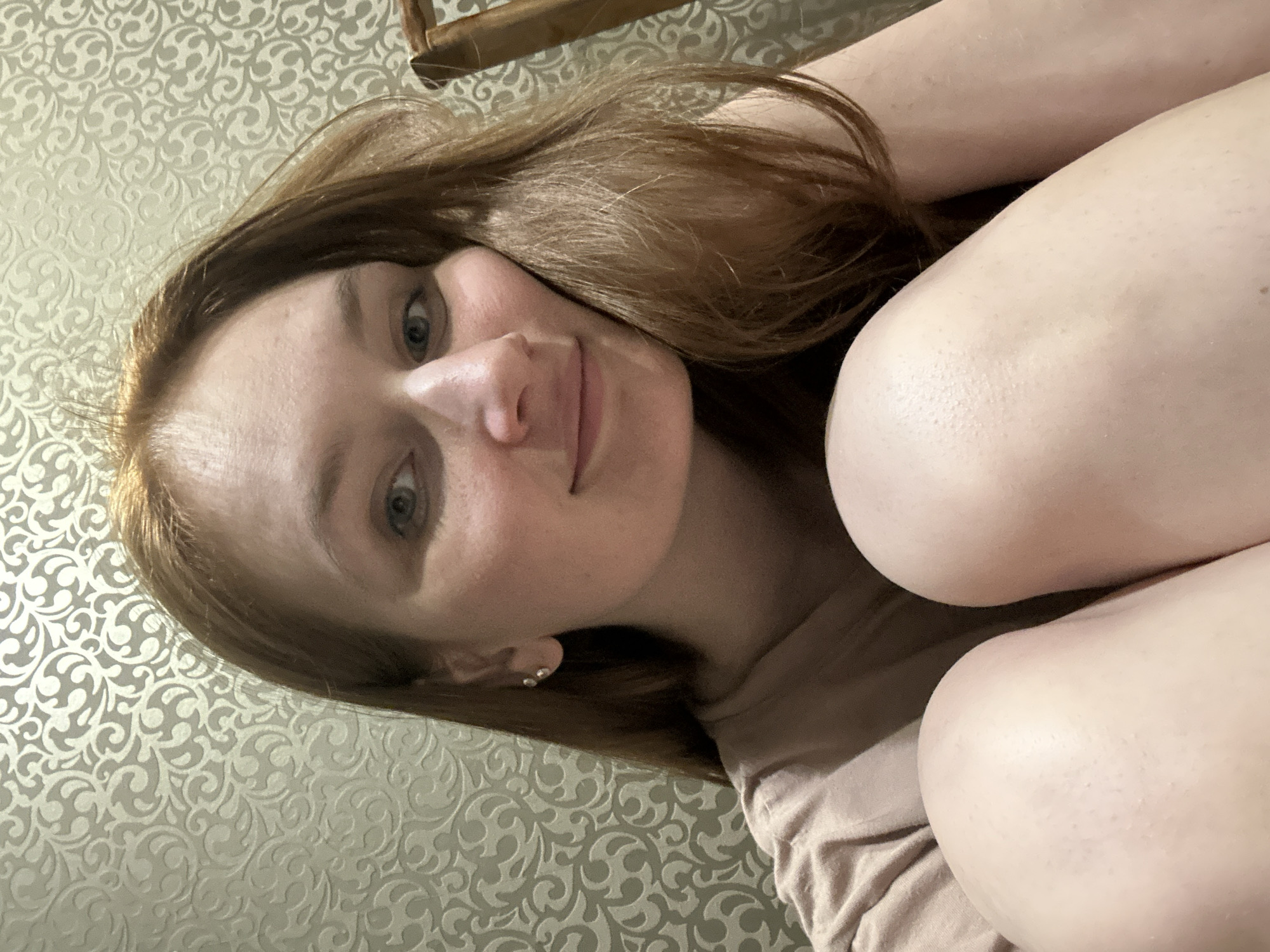A selfie of a fair-skinned woman with auburn hair lying down on a bed with a patterned headboard. The location is plausibly a private residence in Chaikovsky, Russia, given the user's geotagged location. The woman is wearing stud earrings and a light-colored t-shirt. She is looking directly at the camera and smiling slightly. Her knees are drawn up towards her chest. The bedsheets are a similar color to her t-shirt.