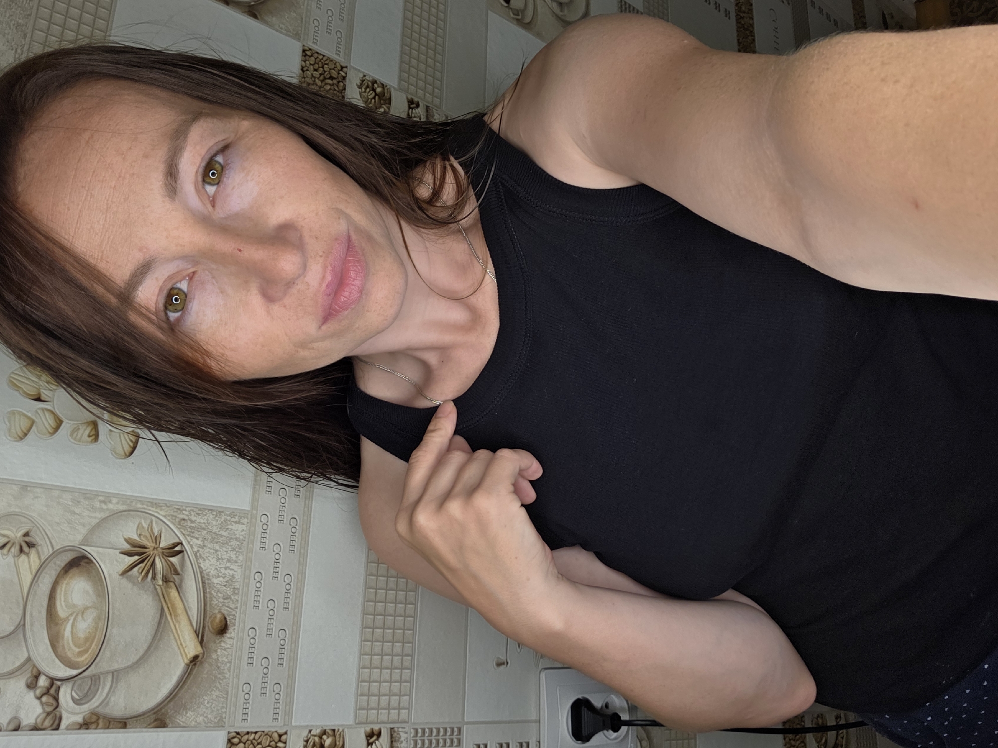 A woman with long brown hair, wearing a black tank top and dark blue polka dot pants, is shown from the chest up. She is indoors, in what appears to be a kitchen based on the wallpaper featuring coffee cups, coffee beans, and tiles. Her right hand is raised, with her index finger pointing towards her collarbone. The background includes a white electrical outlet and cord.