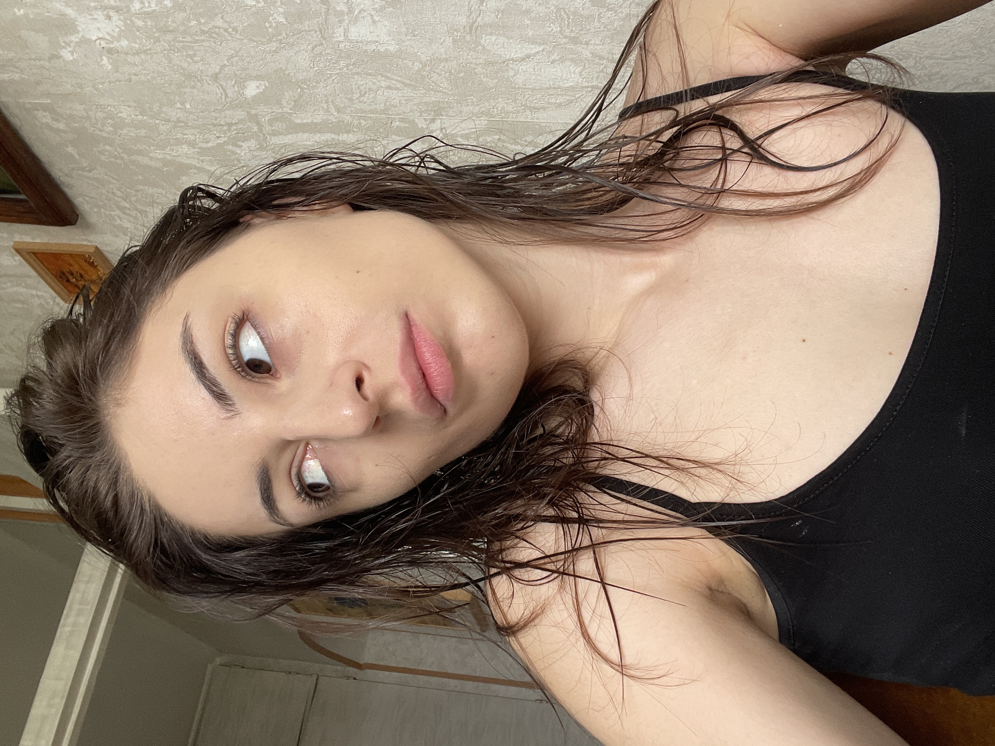 A young woman with dark, wet hair pulled back into a bun is looking upwards and to her left, her head tilted slightly. She is wearing a black tank top. Her skin appears slightly shiny, suggesting it might be damp. The background consists of a textured, light-colored wall and a portion of a doorframe. A small, framed picture hangs on the wall to her upper right. The scene is indoors, in what appears to be a room. There is no information available to determine the specific location within Moscow, Russia.