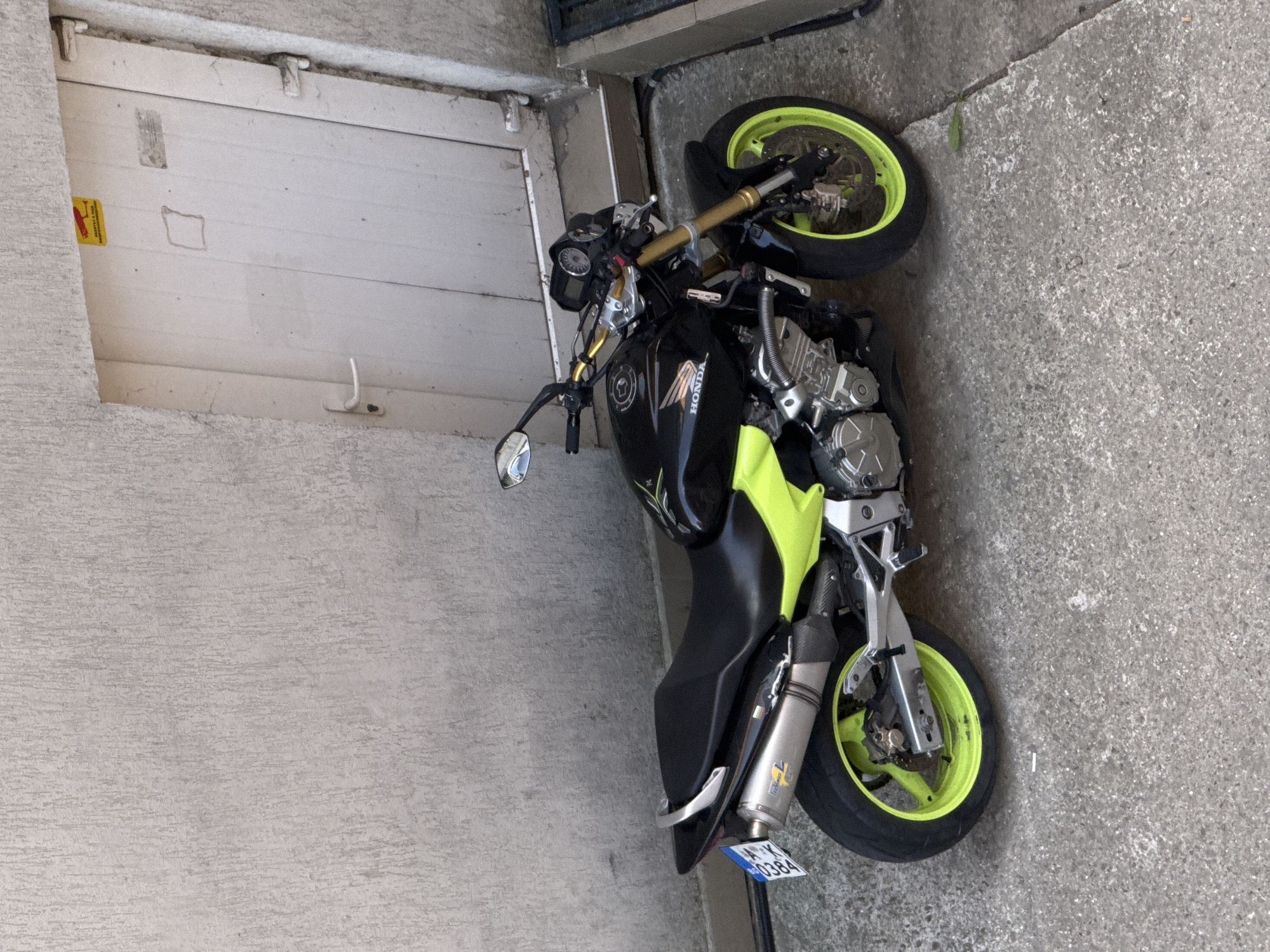 The image shows a black and neon green Honda motorcycle parked in a corner of a building. The motorcycle is parked on a concrete surface next to a light-colored, possibly concrete, wall and a white garage door. The license plate of the motorcycle reads "BHX 0384." The location is most likely Burgas, Bulgaria, based on the license plate.
