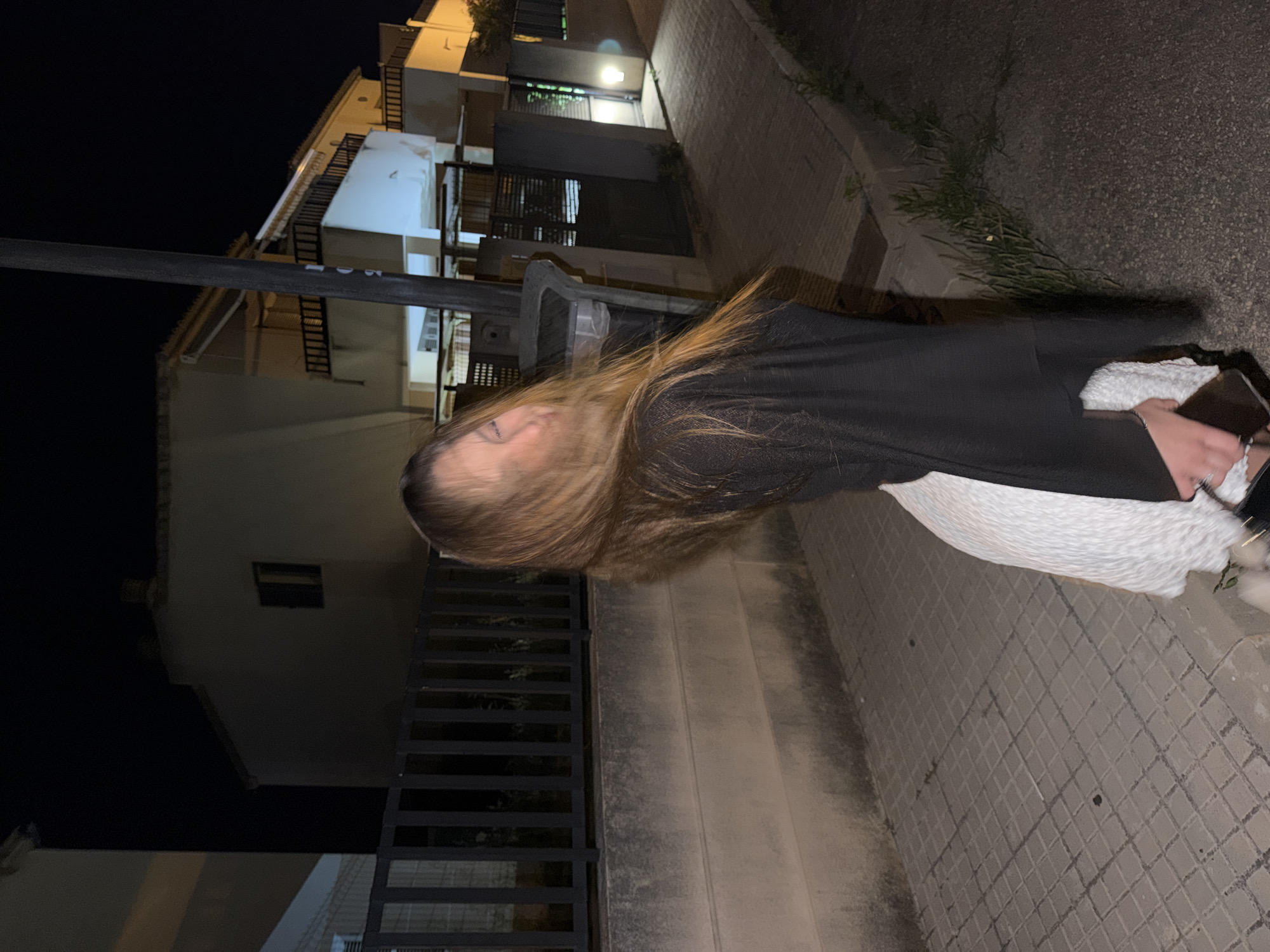 A woman stands on a tiled sidewalk in Platja D'alcúdia, Spain, at night. She wears a black top and a white skirt. She is holding a phone in her right hand. Buildings line the street, illuminated by streetlights. A black metal fence is visible in the foreground.