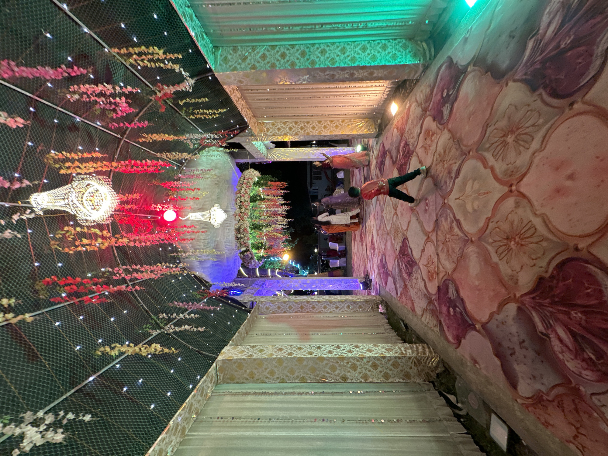 Captured indoors, the image reveals a room lavishly adorned for an event, possibly a wedding, in Shahapur, India. The space features elaborate decorations including flower arrangements, decorative lighting, and patterned fabrics. People dressed in traditional Indian attire are observed within the scene. The perspective of the image suggests it was taken from an elevated position, looking down into the decorated area.