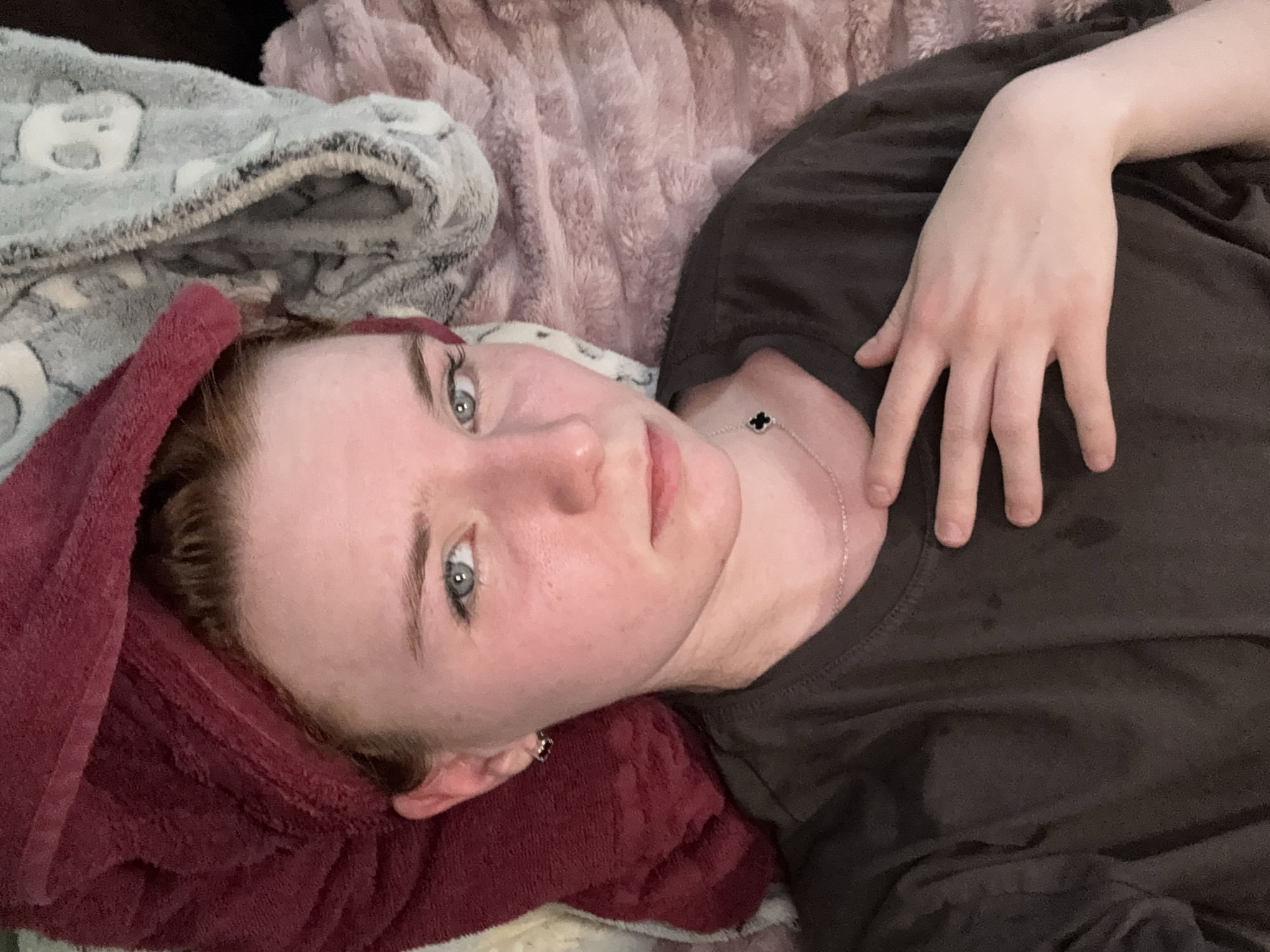 A pale-skinned person with light eyes lies on a bed with a burgundy towel draped over their head. They are wearing a dark-colored t-shirt and a silver necklace with a clover-shaped pendant. Their right hand rests on their chest. The bed is covered with a variety of blankets and plush fabrics in shades of gray and pink. The location is unknown.
