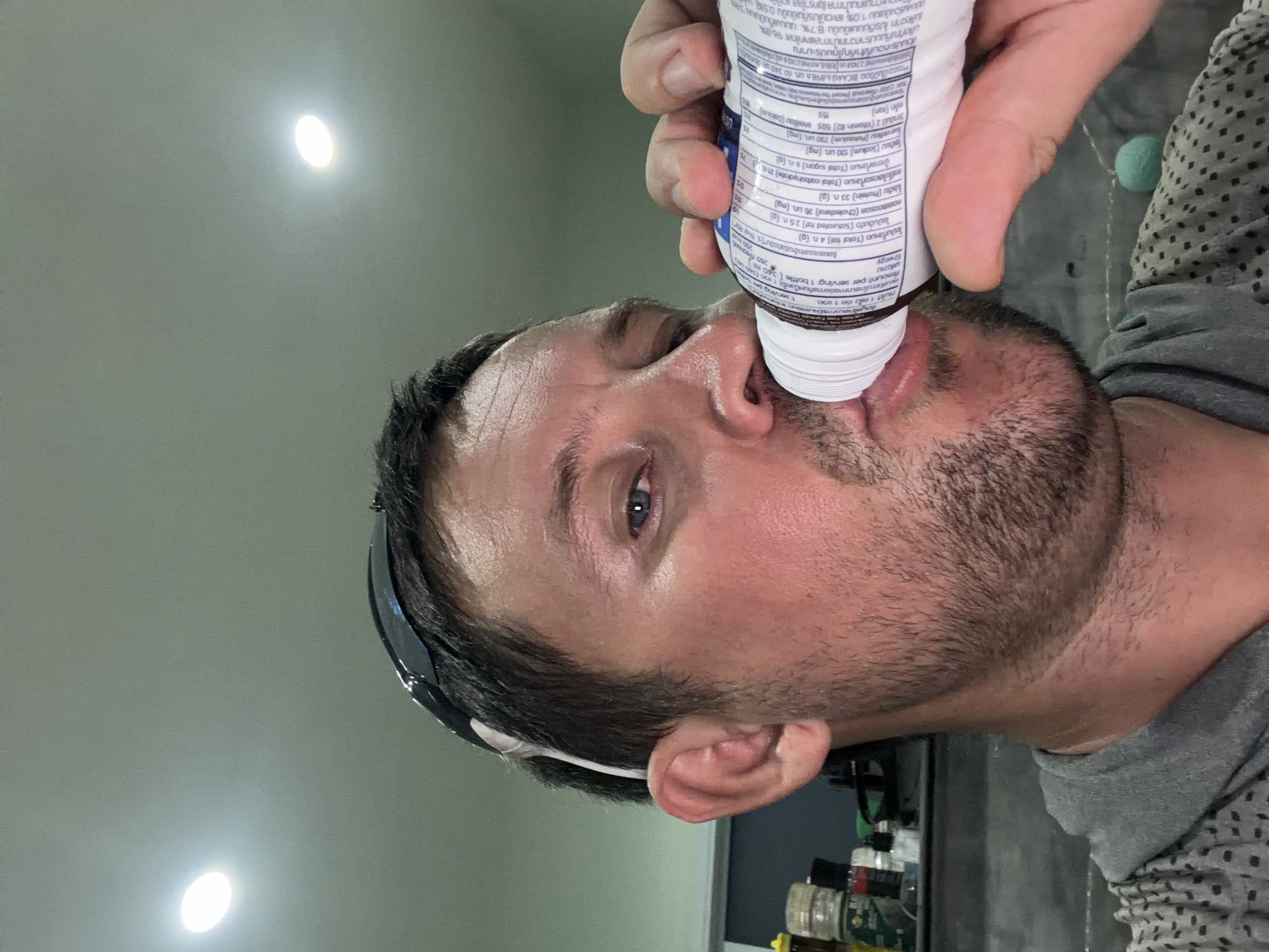 A man is taking a selfie while drinking from a bottle. He is wearing sunglasses pushed up on his forehead. The background appears to be an indoor setting with shelves stocked with various items. The man has short, dark hair and stubble. The lighting suggests artificial light sources from above. The text on the bottle is in a foreign language, likely Thai, indicating the location of Baan Mae Nam, Thailand.