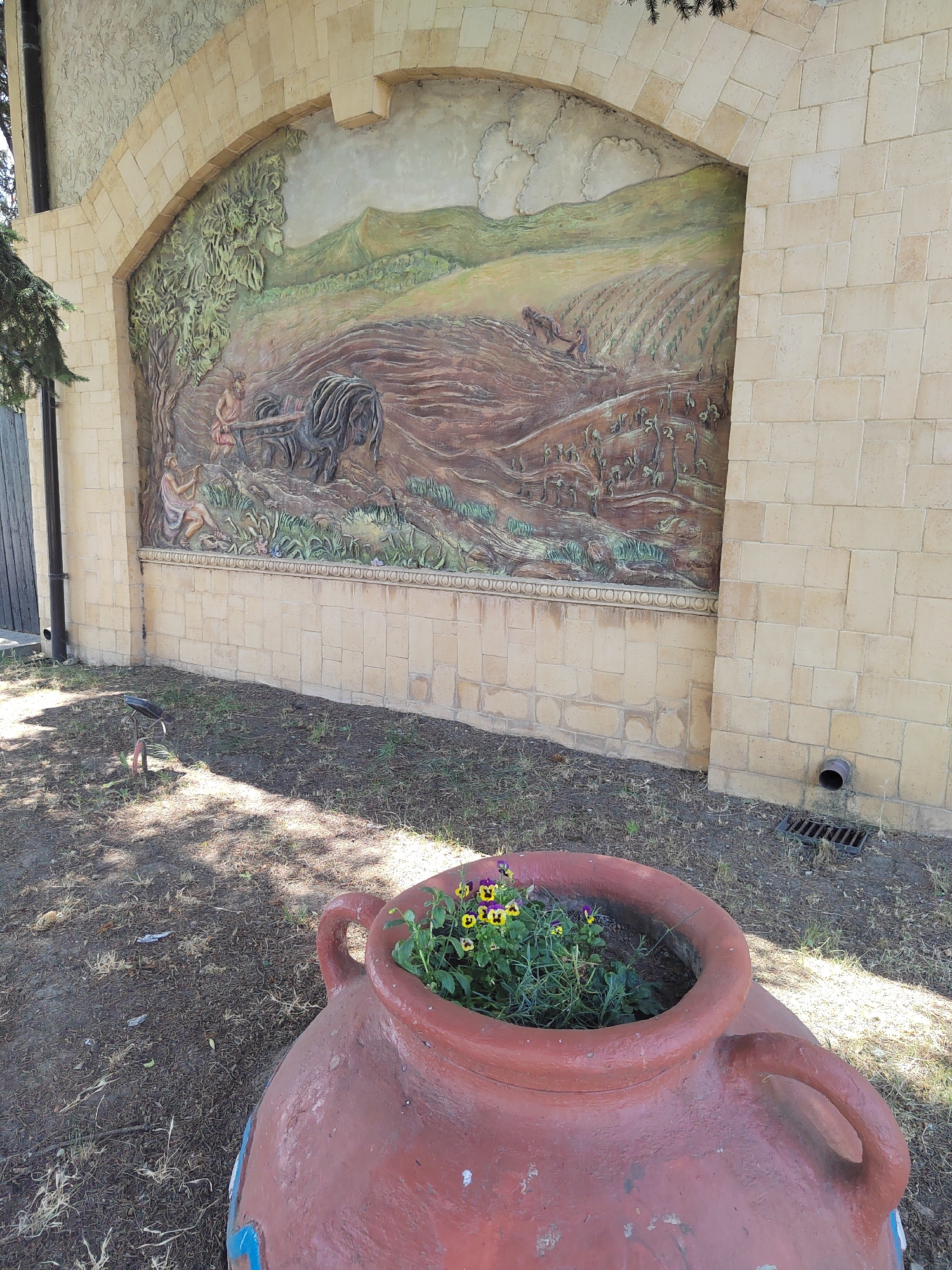 An outdoor scene depicts a large, arched mural on a stone wall. The mural is a bas-relief sculpture depicting an agricultural scene, with figures plowing a field with a horse-drawn plow, and planting crops. The overall color palette of the mural is earthy tones, with greens for foliage and hills, and browns for the soil. In the foreground, a large, red clay pot with handles serves as a planter for small, colorful flowers and greenery. The pot is placed on a patch of dry, uneven ground with sparse grass and shadows cast by nearby trees. To the left of the mural, a tall, dark, vertical pole, possibly a fence post or a lamppost, is visible. To the right of the mural, a drainpipe and a metal grate are embedded in the stone wall at ground level. The stone wall is constructed from light-colored, rectangular blocks. The mural is situated in Vityazevo, Russia.