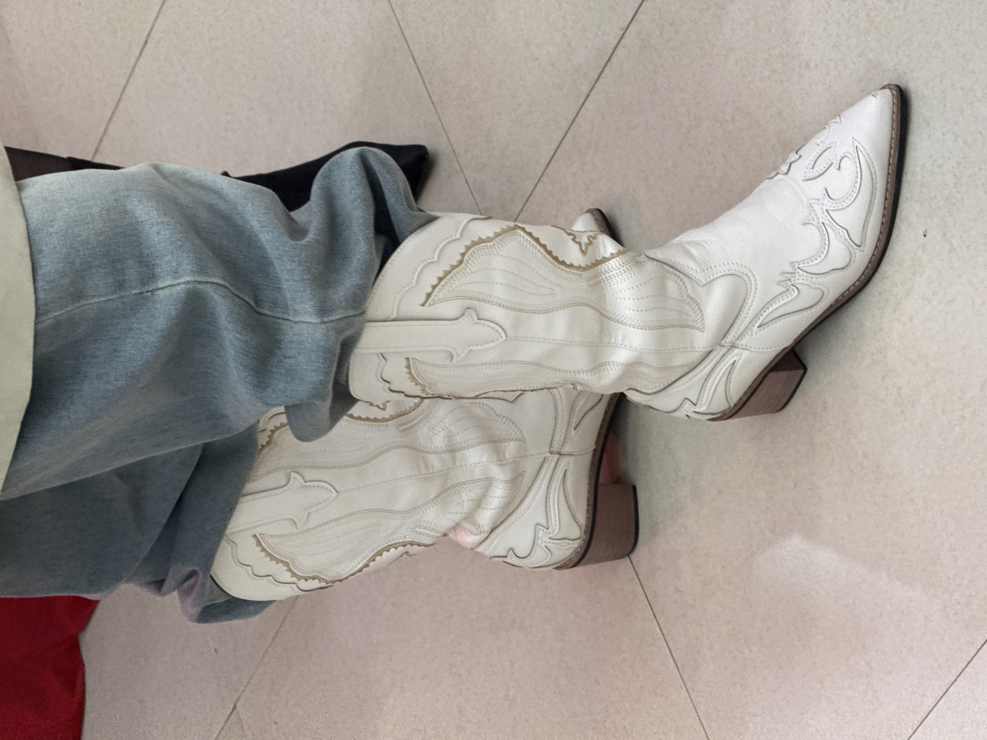 The image shows a person wearing white cowboy boots with decorative stitching. The person is also wearing light blue denim jeans. The boots and jeans are on a light-colored tiled floor. Other clothing items, including a red garment, are partially visible. The location is not determinable from this image alone.